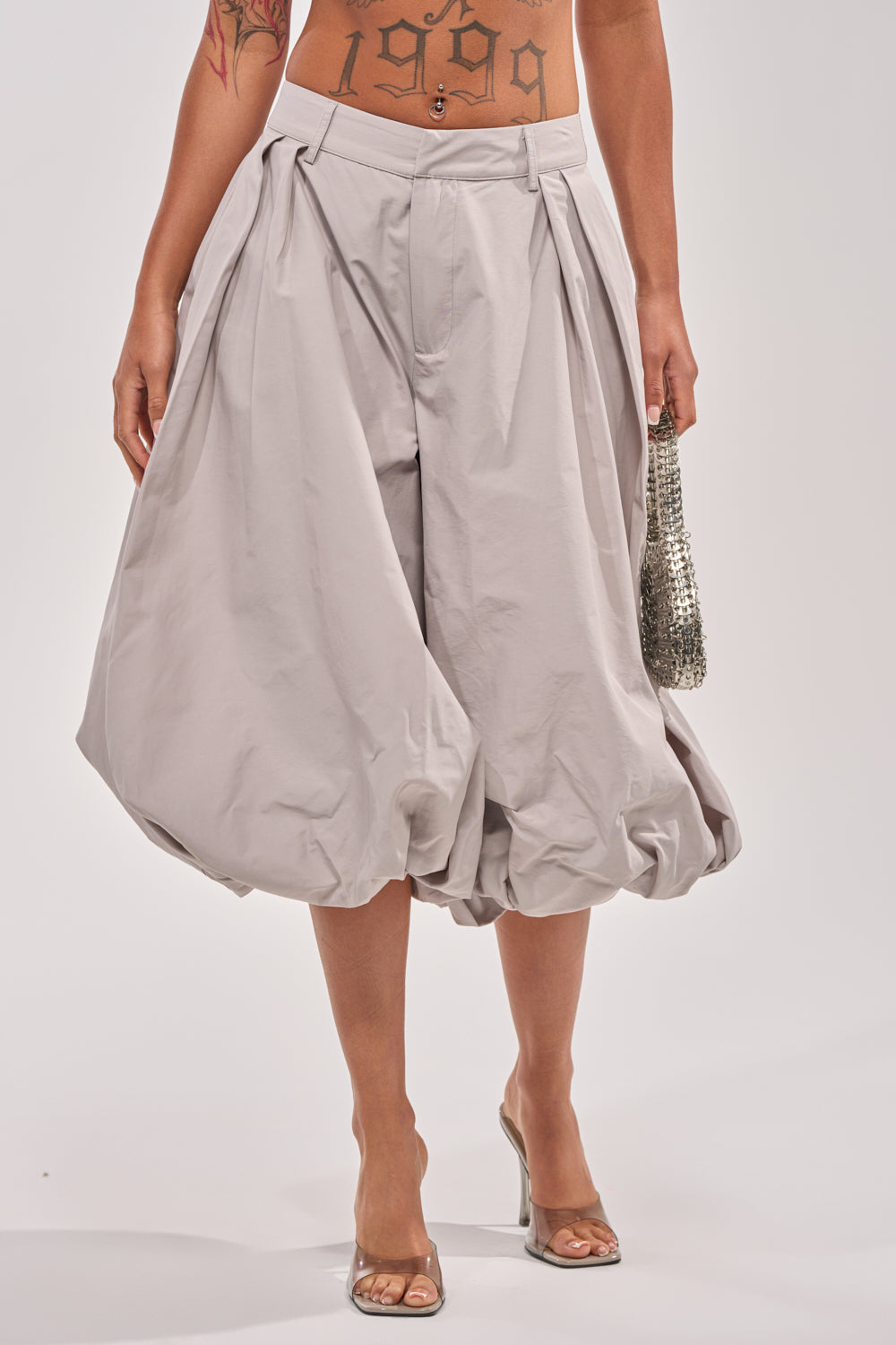 A person wearing GLOW GAUCHO PANT in grey with pleated detailing, clear heeled shoes, and a small silver bag. Only the lower torso and legs are visible; a "1999" tattoo appears on the lower stomach—ideal for contemporary daywear. 2