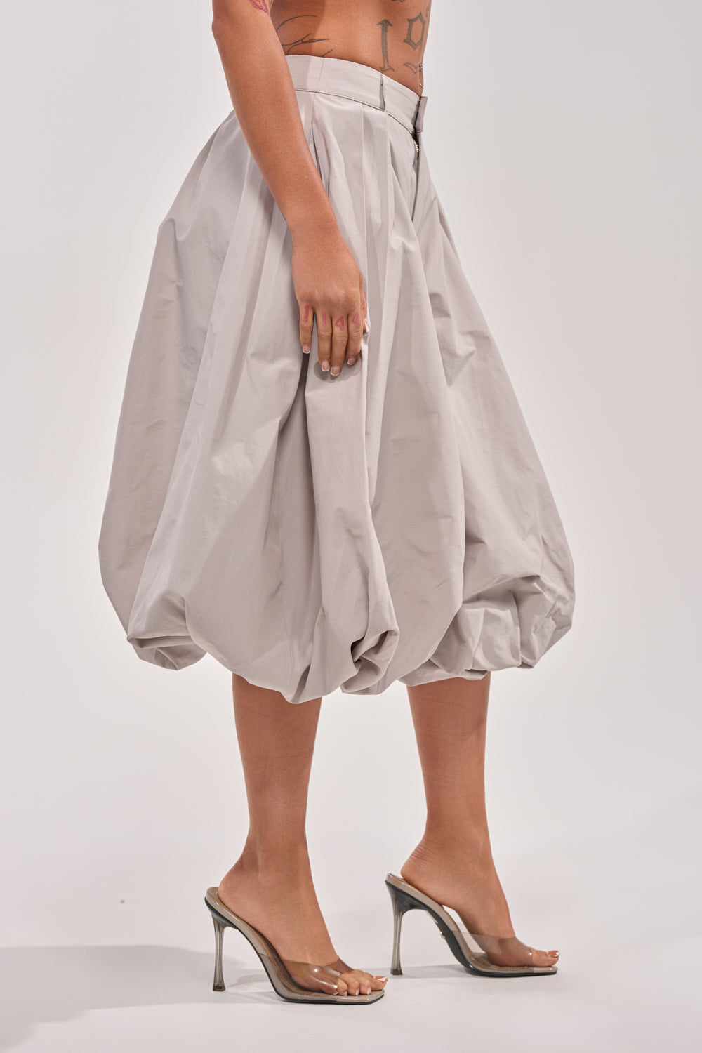 A person models the GLOW GAUCHO PANT in gray with clear high-heeled shoes, standing against a plain light background. Only their torso and legs are visible, highlighting the pant's contemporary, voluminous silhouette. 3