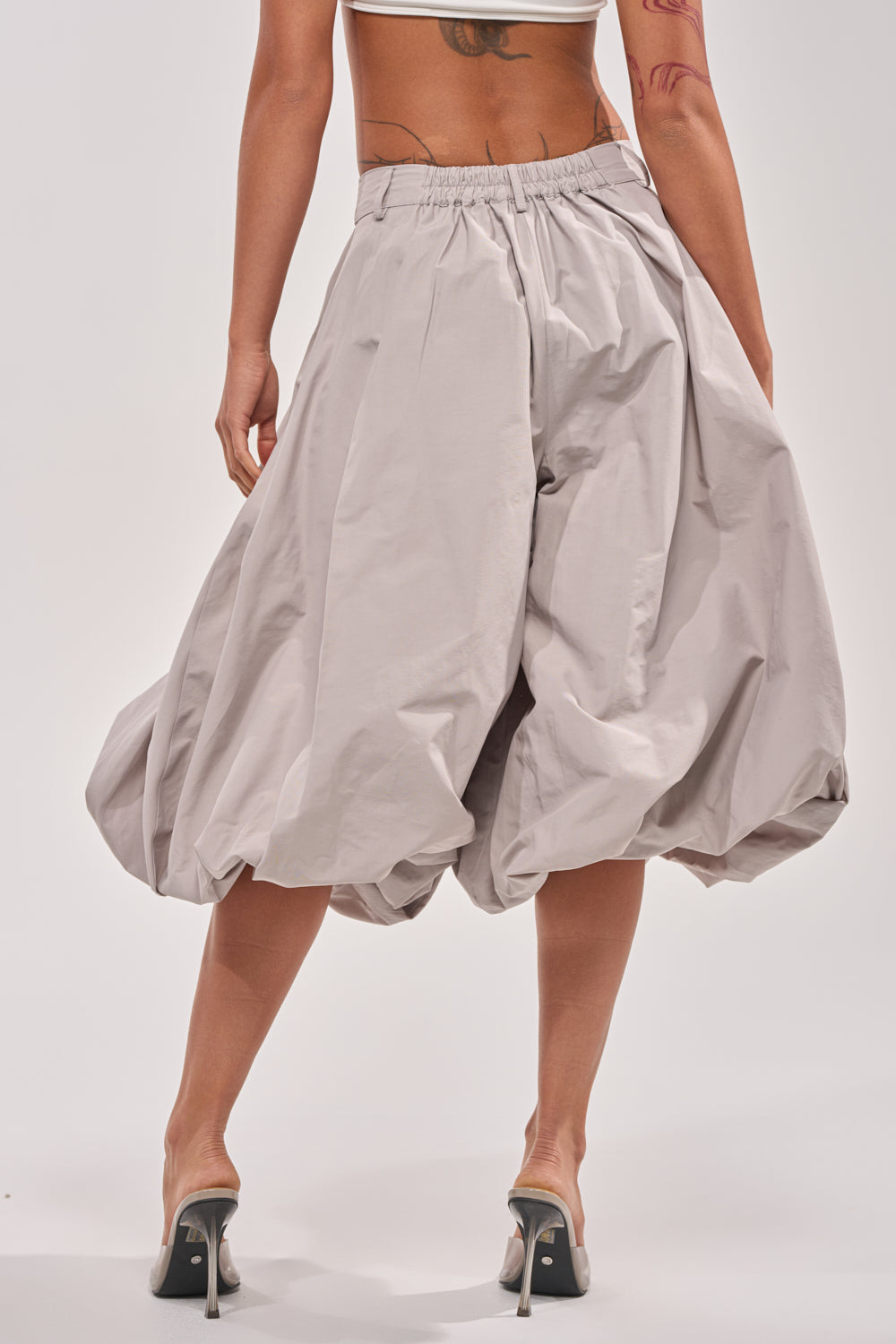 A person in modern style wears GLOW GAUCHO PANT in grey with voluminous legs, a white crop top, and high-heeled sandals, standing back to the camera to display tattoos on their back and arm. 4