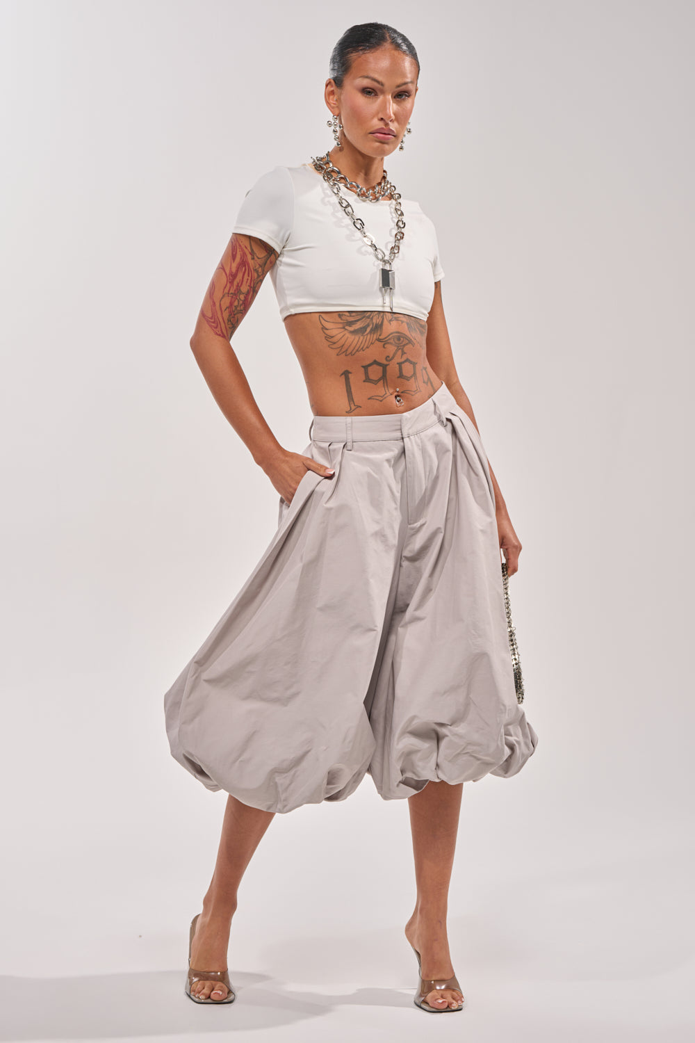 Model showcases the GLOW GAUCHO PANT in grey with a white crop top and heeled sandals. Hoop earrings, tattoos, and a chunky chain necklace add edge to this confident, modern look against a plain backdrop. 5