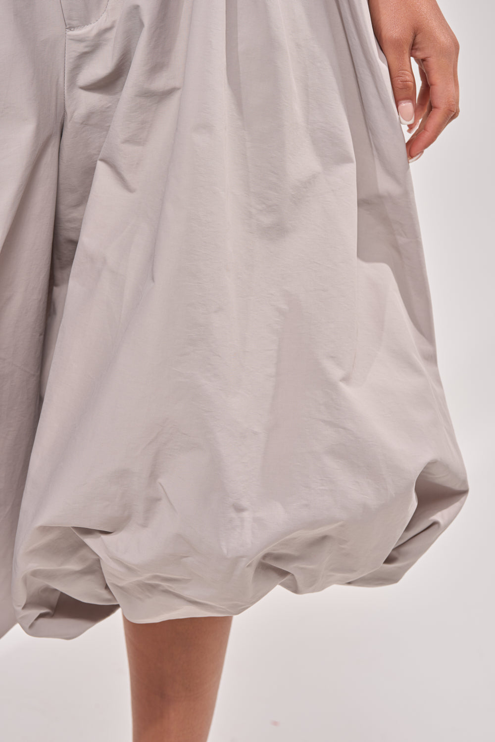 A close-up of a hand resting by the side, wearing contemporary daywear—the GLOW GAUCHO PANT in light grey with a voluminous, balloon-style hem—set against a plain light background. 6