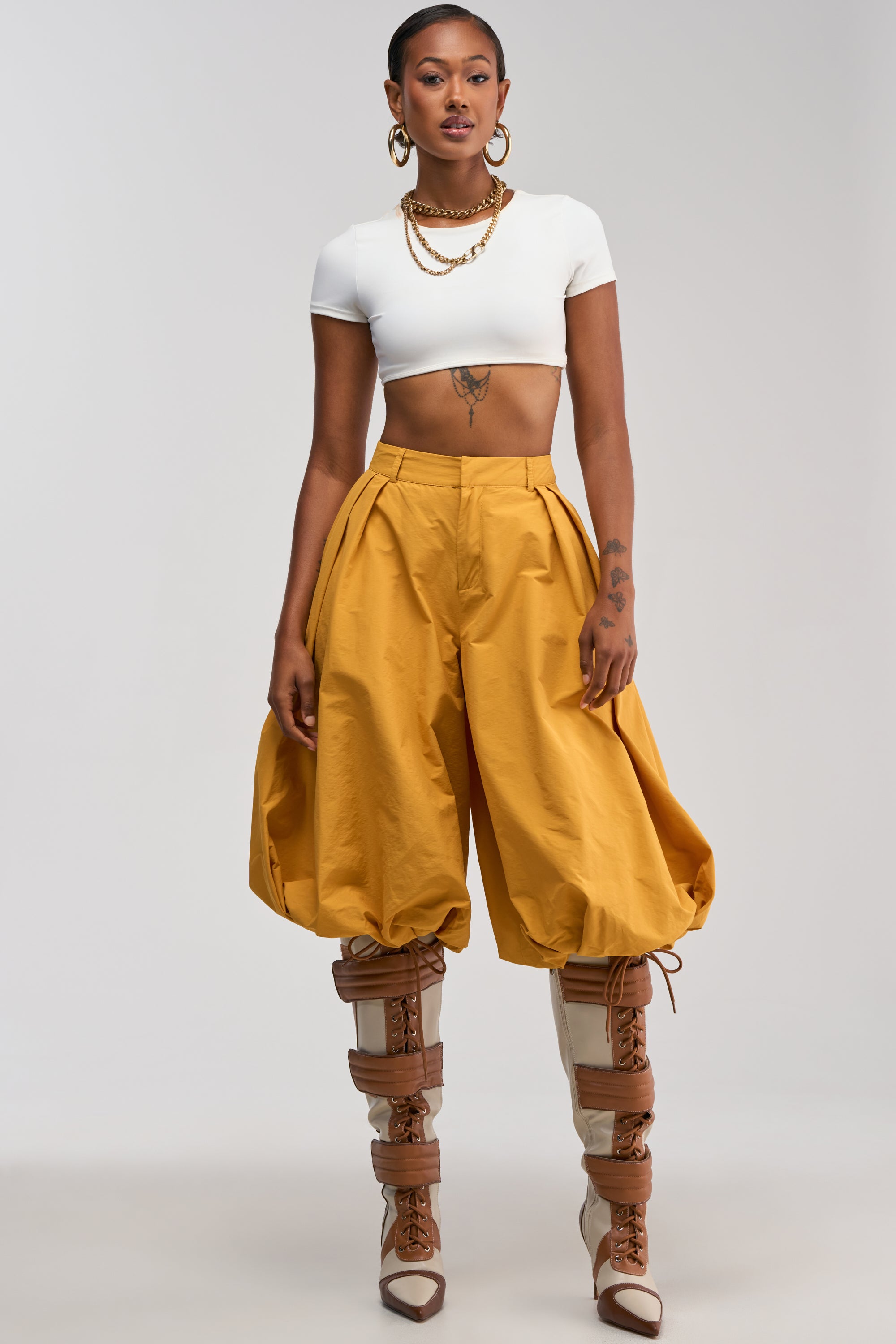 A woman stands facing forward against a plain light background, wearing the GLOW GAUCHO PANT IN MUSTARD styled with a white crop top, tall lace-up boots, large hoop earrings, and layered gold necklaces.