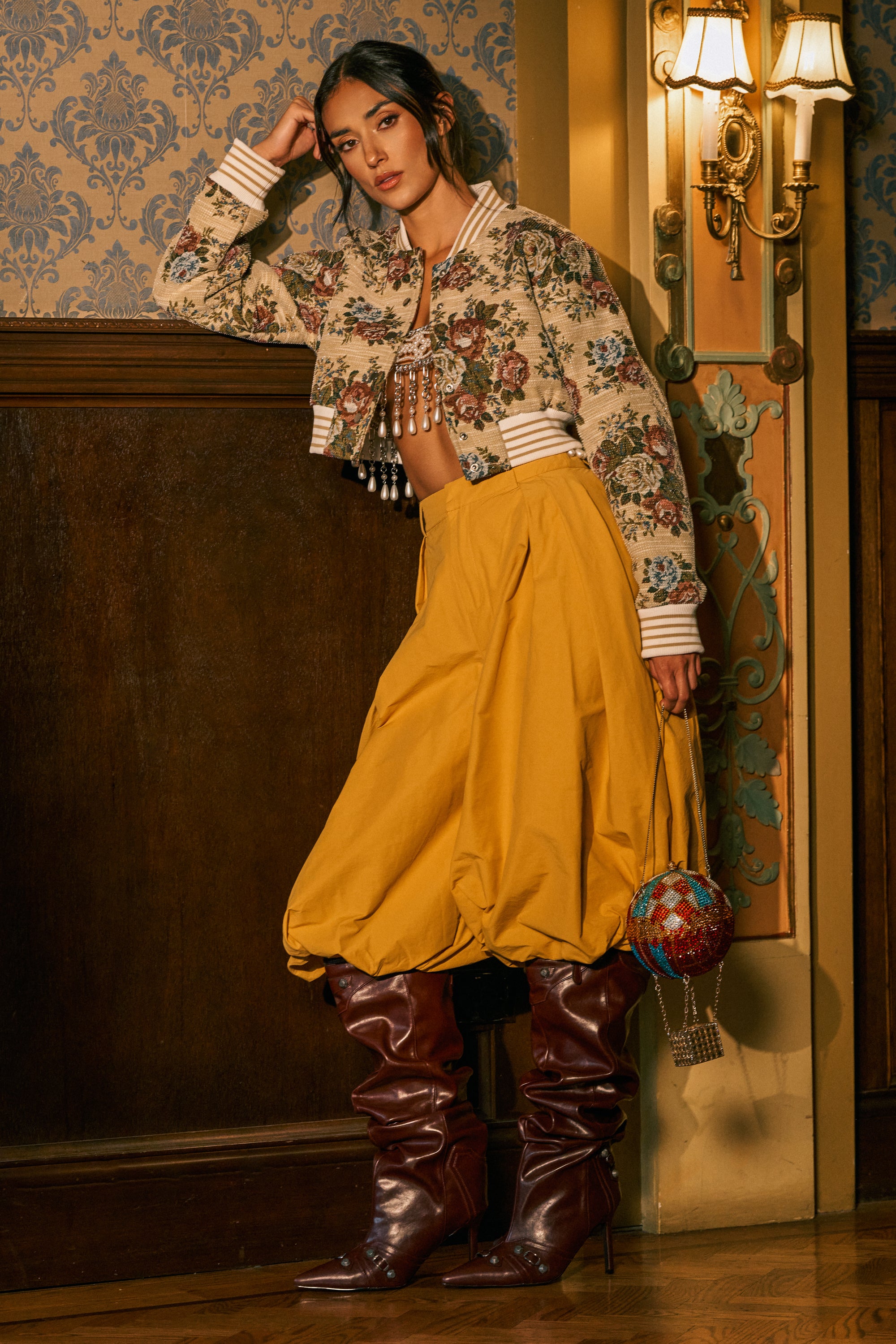 A woman poses indoors wearing the GLOW GAUCHO PANT IN MUSTARD with a balloon leg silhouette, paired with a floral embroidered jacket, tall brown boots, and holding a colorful beaded handbag, set against ornate wallpaper and wooden paneling.