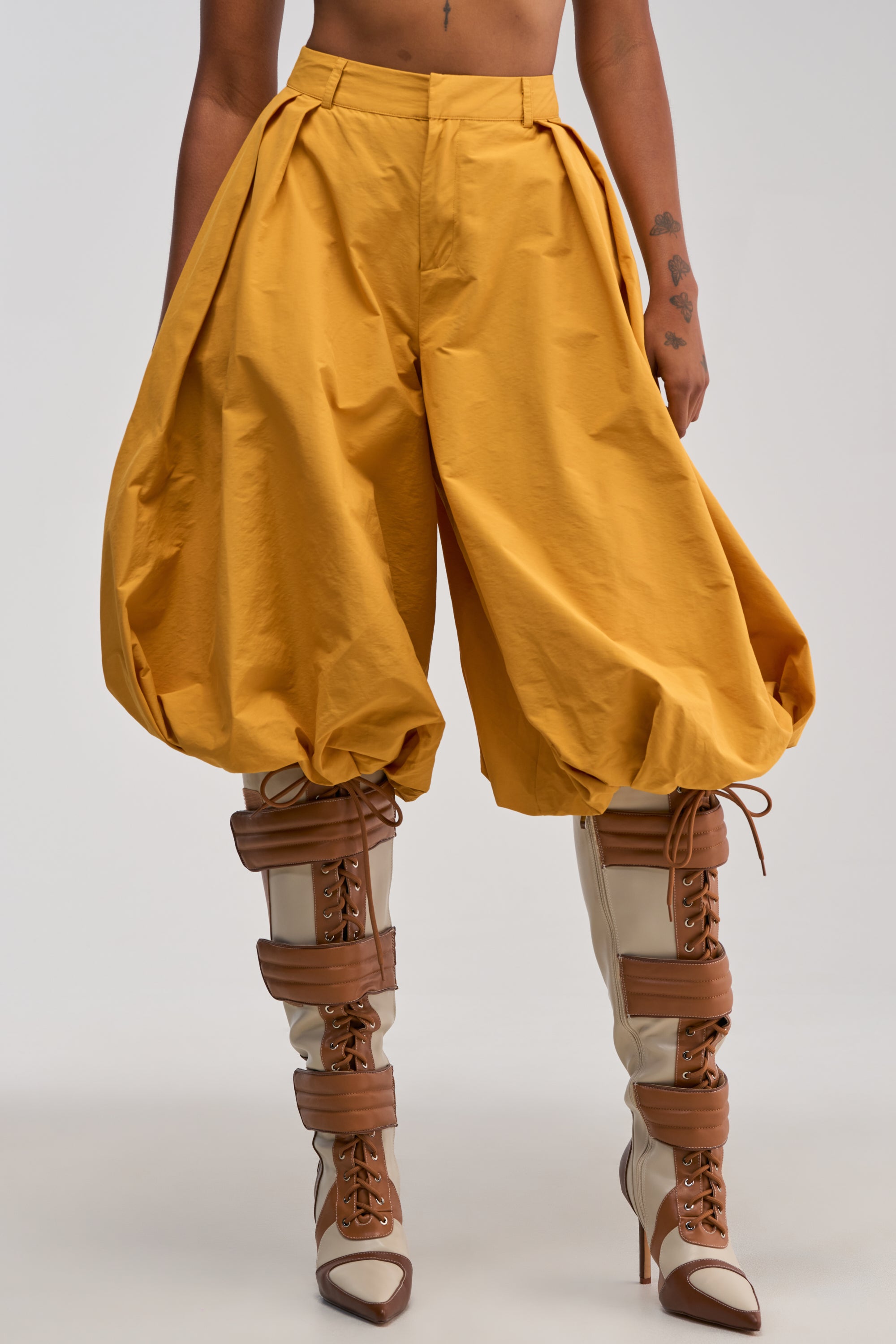A person wears the GLOW GAUCHO PANT IN MUSTARD, featuring a distinctive balloon leg silhouette gathered at the ankles, and pairs them with two-tone brown and cream knee-high lace-up boots while standing against a plain background.