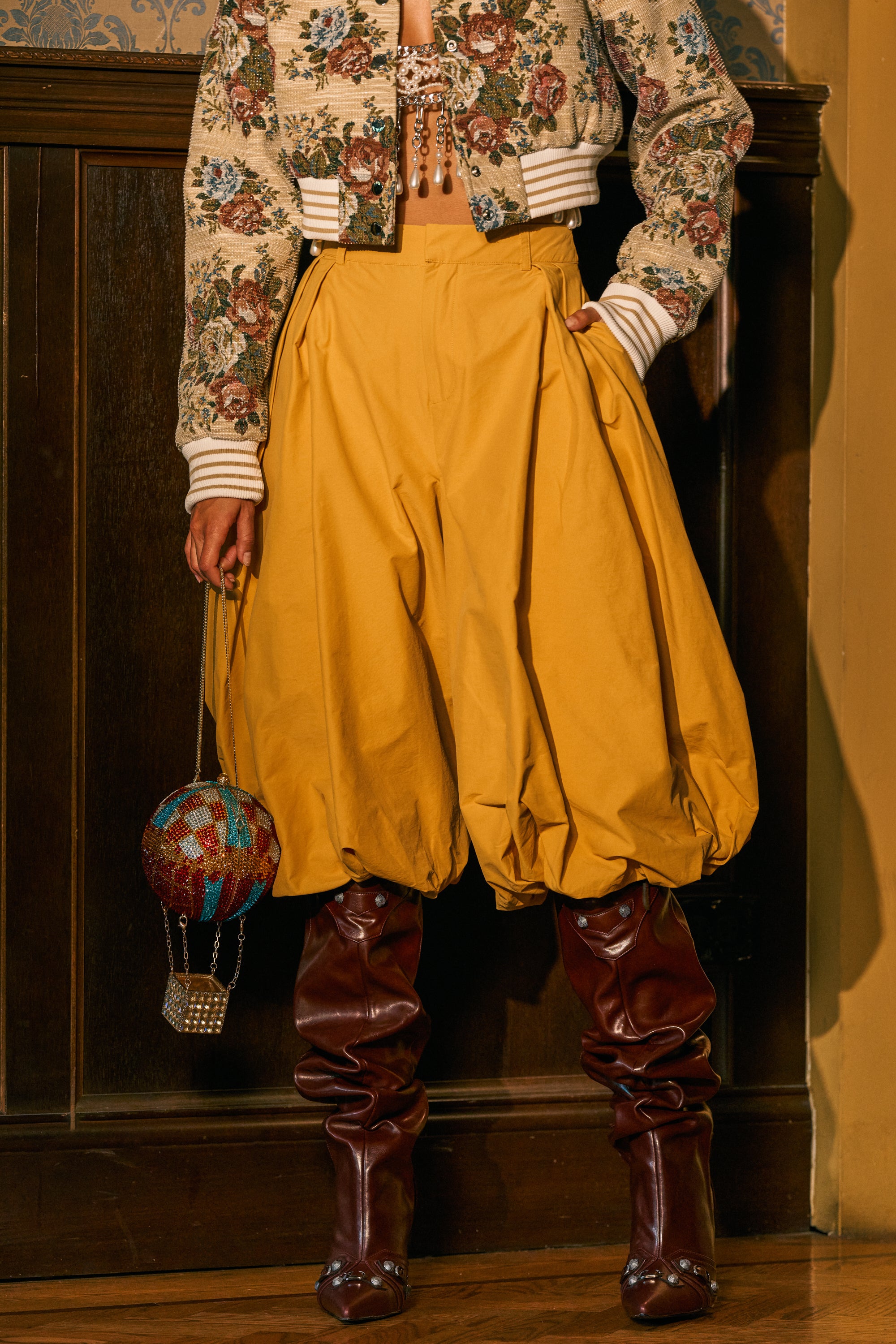 Wearing the GLOW GAUCHO PANT IN MUSTARD, a floral cropped jacket, slouchy burgundy boots, and carrying a beaded hot air balloon-shaped handbag, a person stands against a wood-paneled wall, effortlessly showcasing streetwear style.