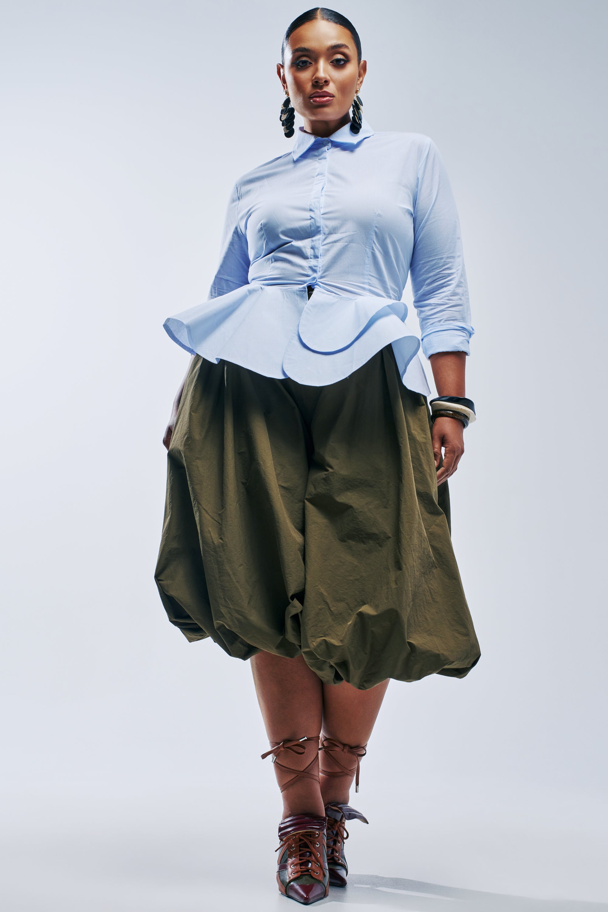 A woman wears a light blue button-up peplum shirt and voluminous, high-rise olive GLOW GAUCHO PANT, paired with lace-up heeled sandals.