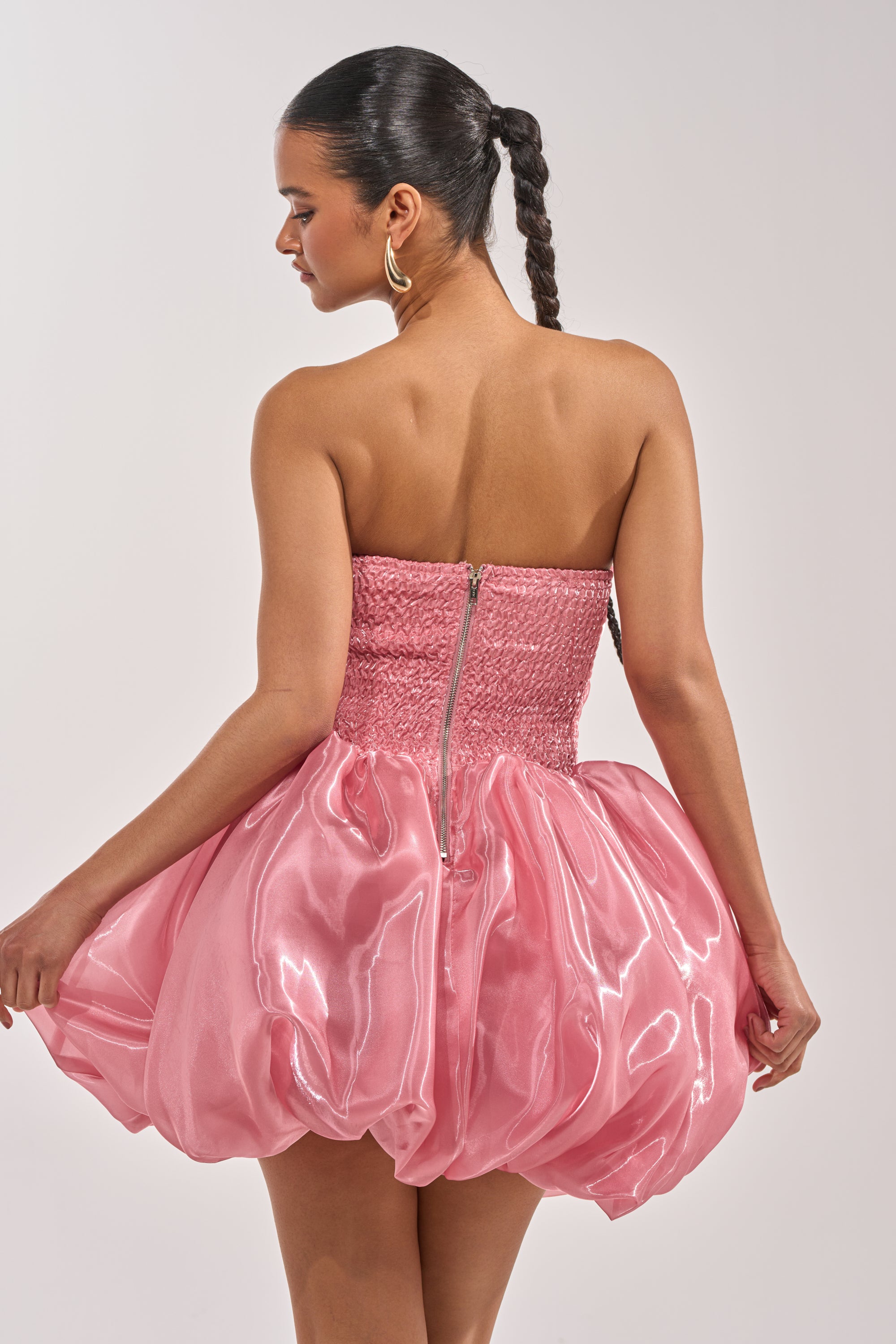 Woman with a sleek braided ponytail and gold hoop earrings stands back to camera in the HARD TO PLEASE BUBBLE MINI DRESS—a shiny, strapless organza mini with sweetheart neckline, smocked bodice, bubble hem, and visible zipper. 3
