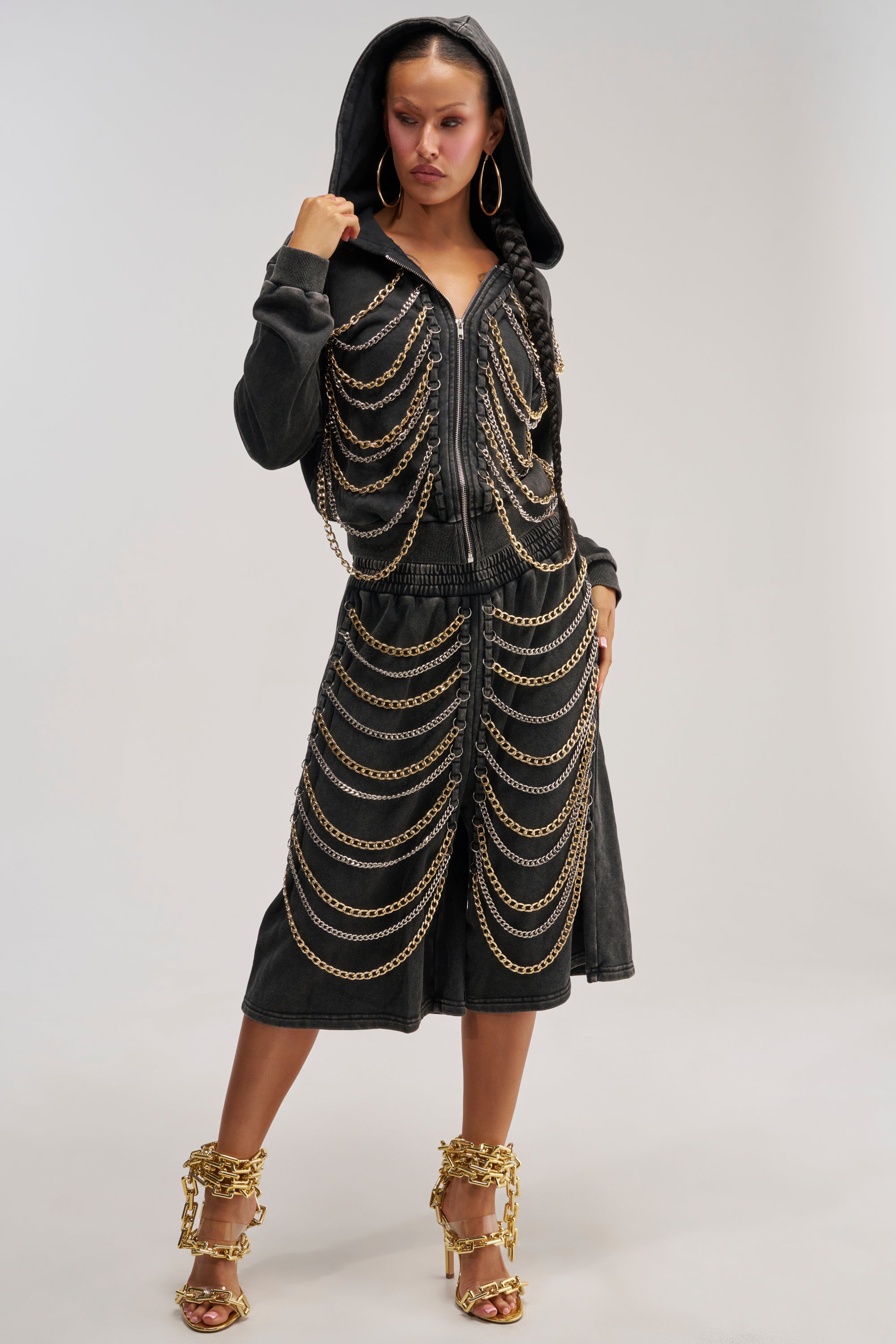 Wearing the CHAINED DOWN BERMUDA SHORT, a woman stands confidently with one hand on her hip, styled in a black hooded jacket featuring striking chain details. She completes the look with gold hoop earrings and gold chain-embellished high heels.