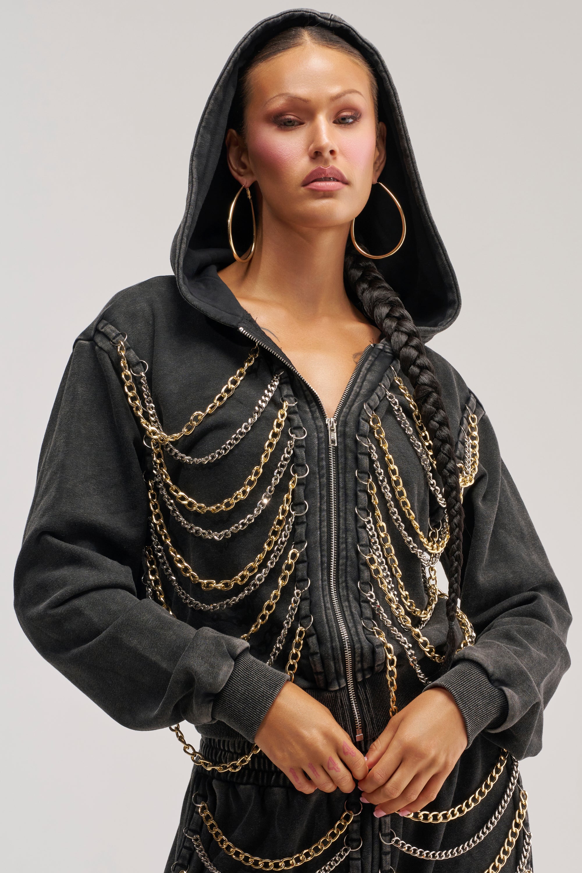 A woman wears the CHAINED DOWN SWEATSHIRT in washed black, featuring statement chain detailing. She completes her bold streetwear look with multiple gold chains, large hoop earrings, and a long braided hairstyle while standing against a plain background.