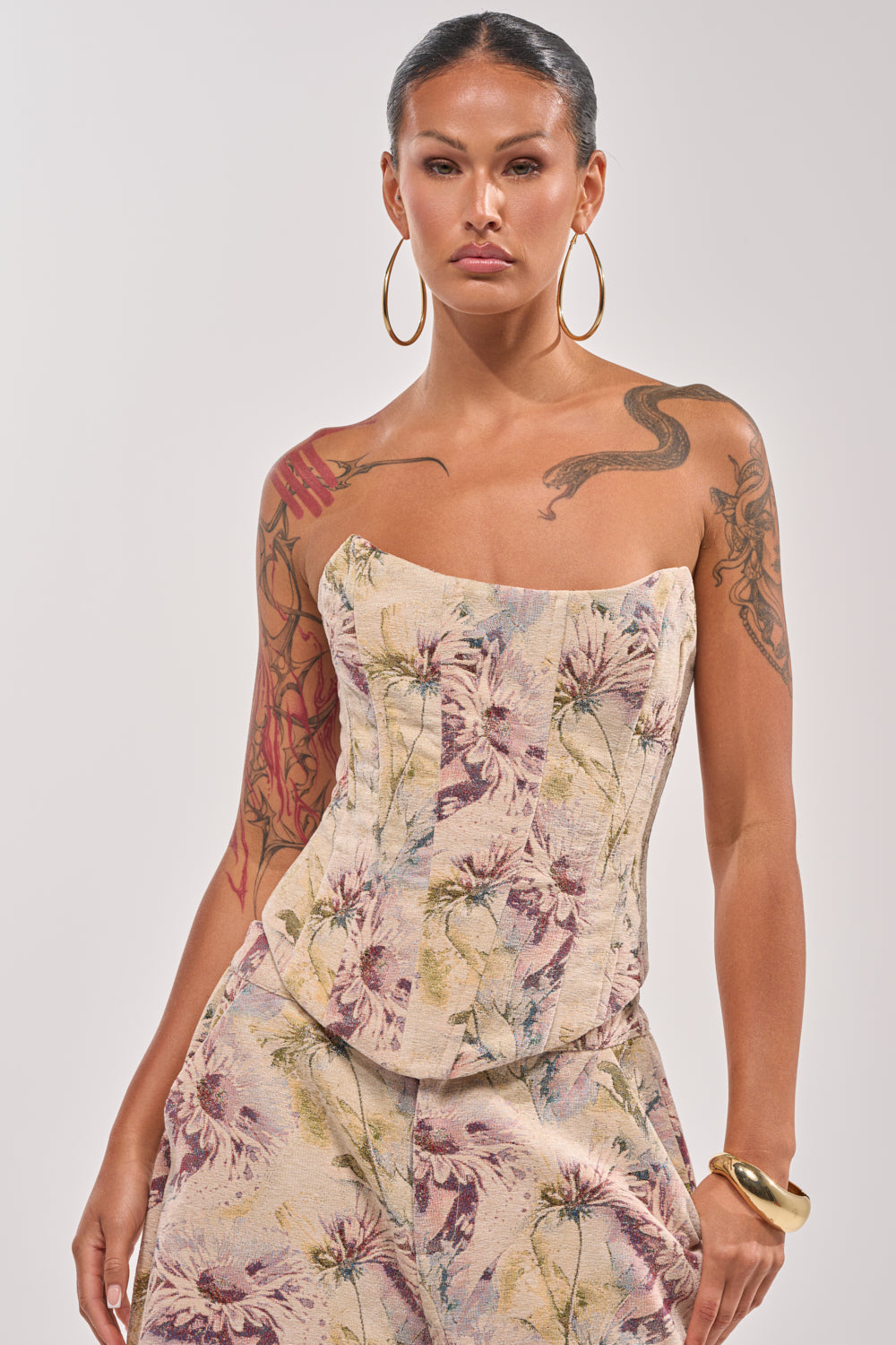 A woman with large hoop earrings and tattoos poses confidently in the SWEETIE PIE BROCADE CORSET TOP and matching pants, her slicked-back hair and chunky right bracelet completing the look of a romantic evening outfit. 1