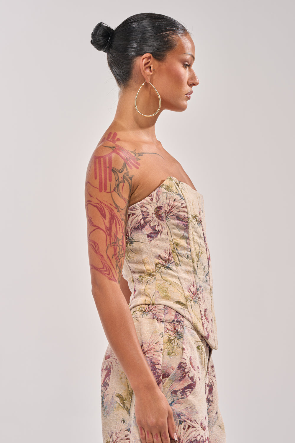 A woman with her hair in a bun and large hoop earrings stands in profile wearing the SWEETIE PIE BROCADE CORSET TOP in patterned beige, showing off a vibrant floral tattoo sleeve on her upper arm and shoulder. 2