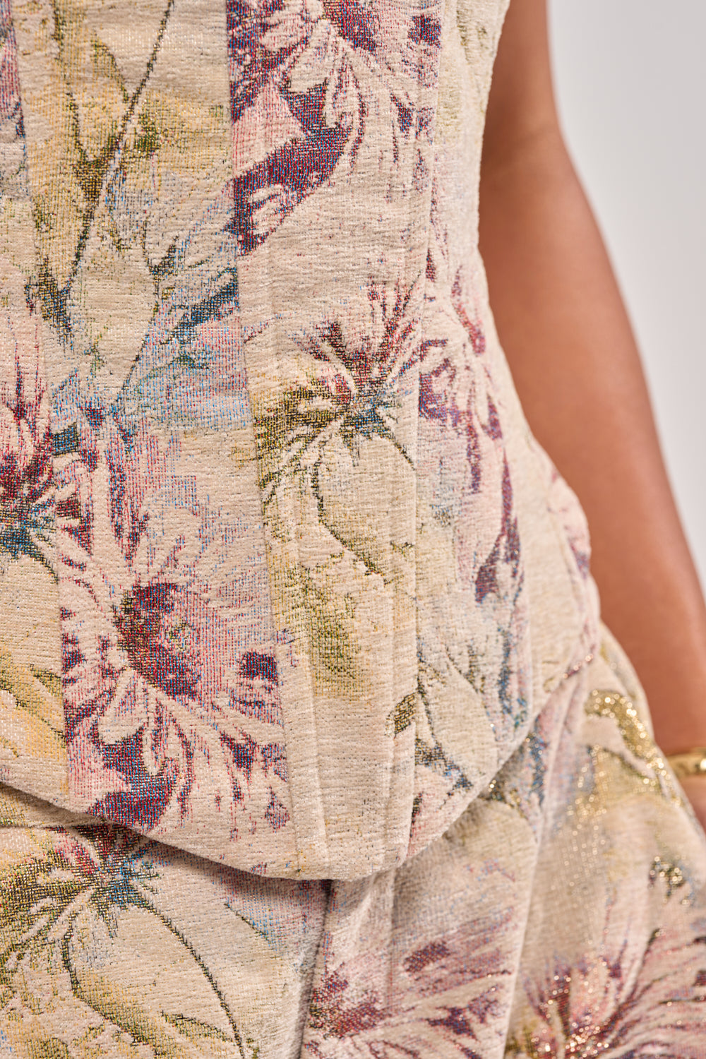 Close-up of a person wearing the SWEETIE PIE BROCADE CORSET TOP in muted purple, green, and gold. Paired with a textured beige outfit, their right arm is adorned with a gold bracelet. 6