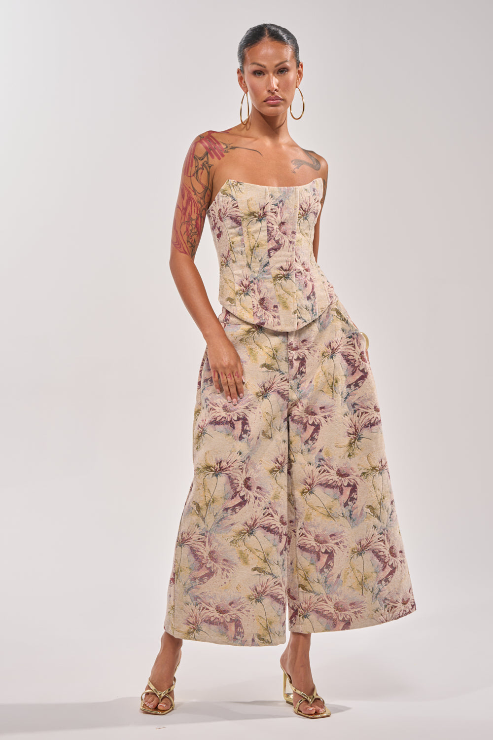 A woman poses confidently in heeled sandals and hoop earrings, showing arm tattoos, while wearing the SWEETIE PIE BROCADE PANT—a wide-leg, cropped pant with a pale botanical print—paired with a strapless fitted top against a plain background. 0