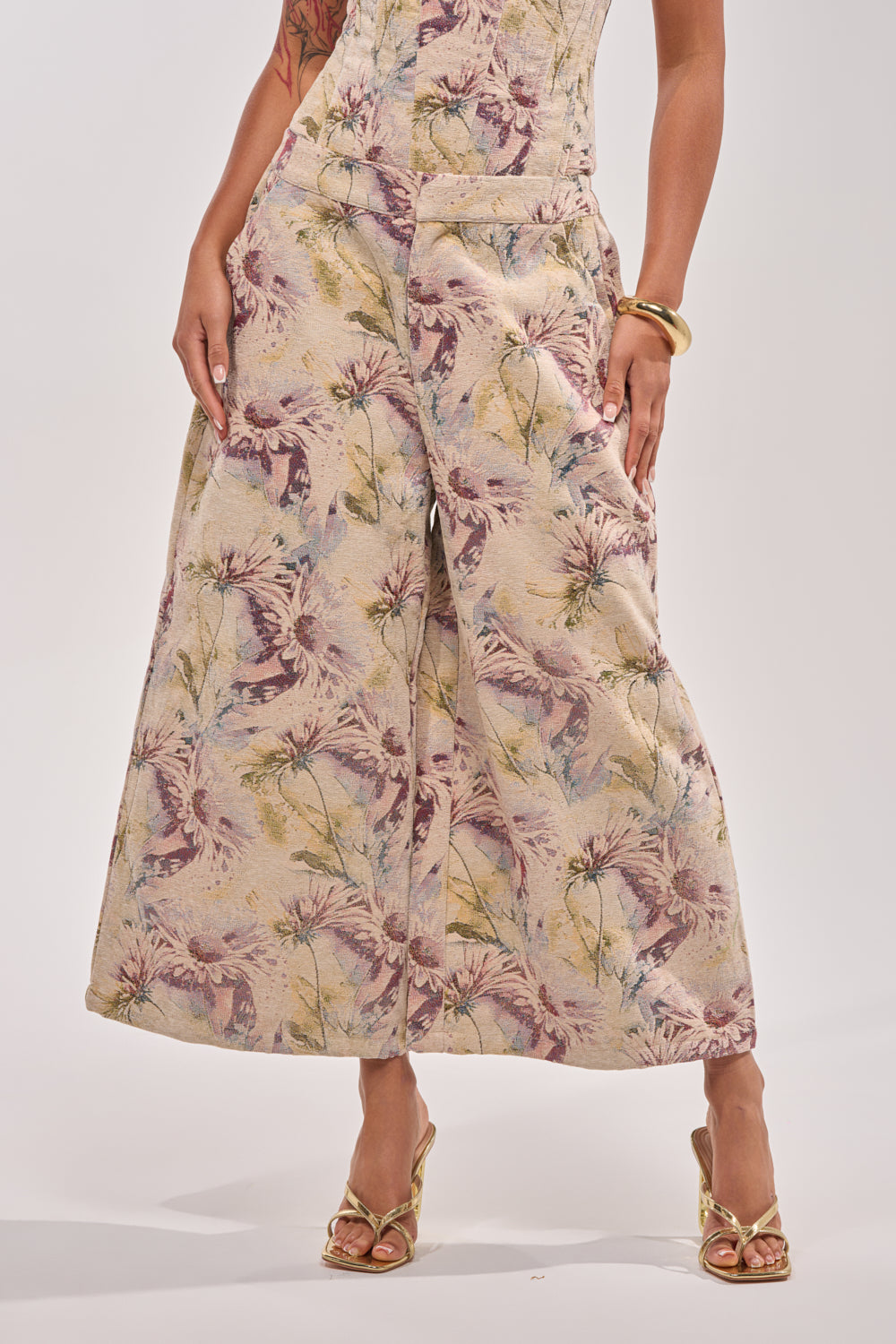 A person wears the SWEETIE PIE BROCADE PANT—a wide-leg, high-rise pant with an elastic waistband in pastel floral light beige—paired with gold sandals and a bracelet, posed against a plain white background. 1