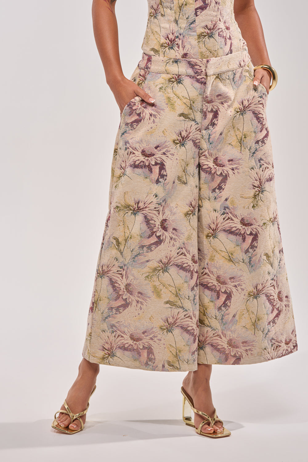 A person stands against a plain light background wearing the SWEETIE PIE BROCADE PANT—floral-print, wide-leg cropped pants with a high-rise elastic waistband—styled with gold open-toe heels and a gold bracelet, hands in pockets. 2