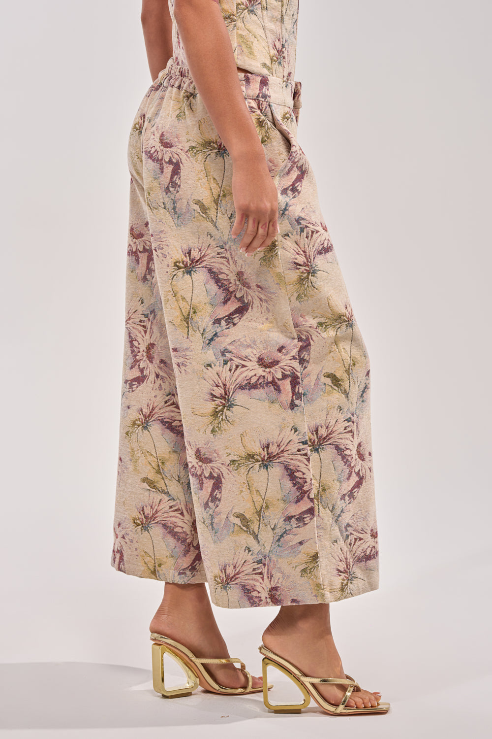 A model stands sideways wearing the SWEETIE PIE BROCADE PANT—wide-leg, cropped, with a pastel floral pattern—and beige strappy heeled sandals. The upper body and face are out of frame against a plain background. 3