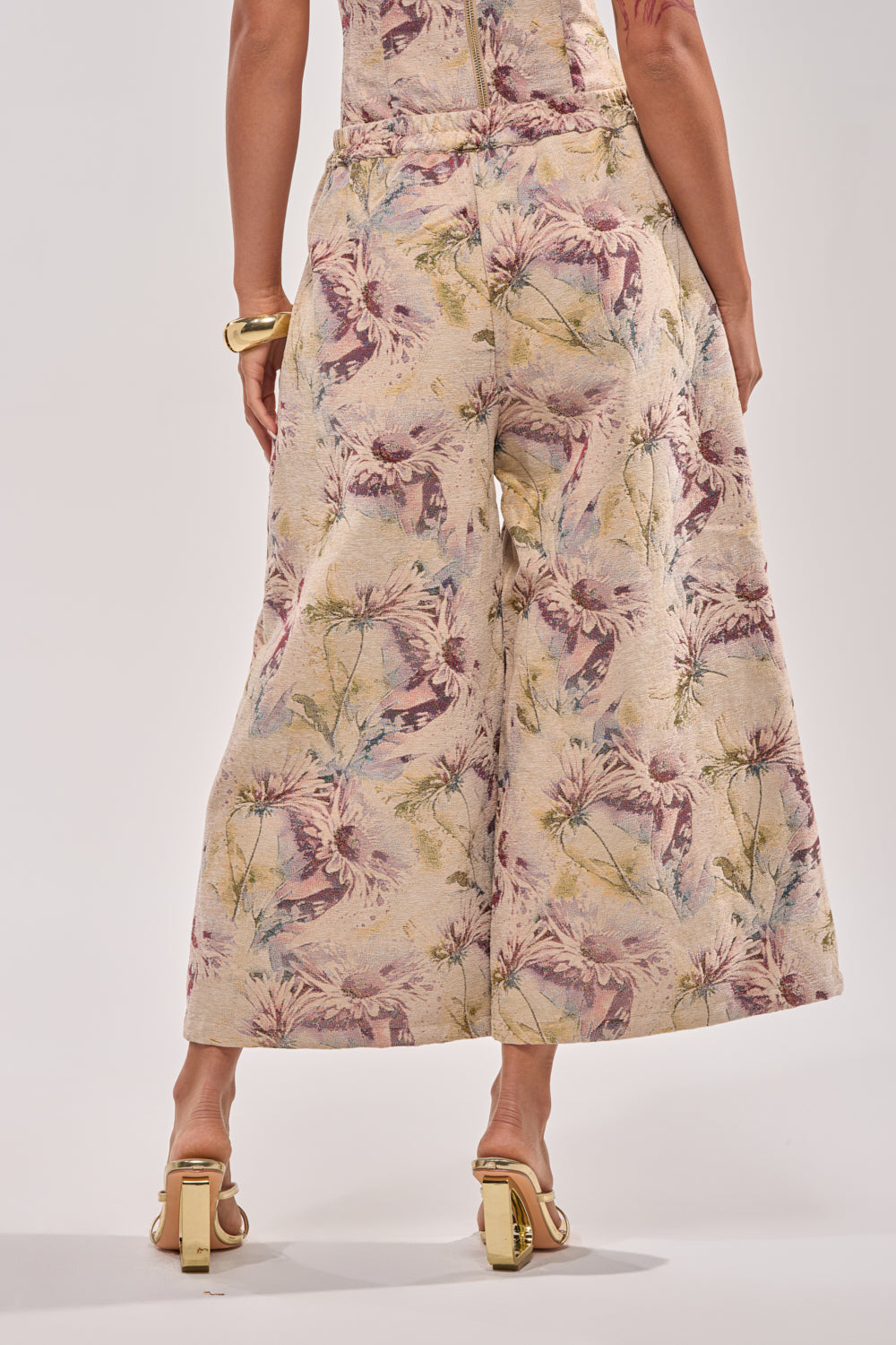 A person stands facing away, wearing the SWEETIE PIE BROCADE PANT—wide-leg, cropped with a floral brocade and high-rise elastic waist—paired with gold heeled sandals and bracelet, against a neutral light background. 4