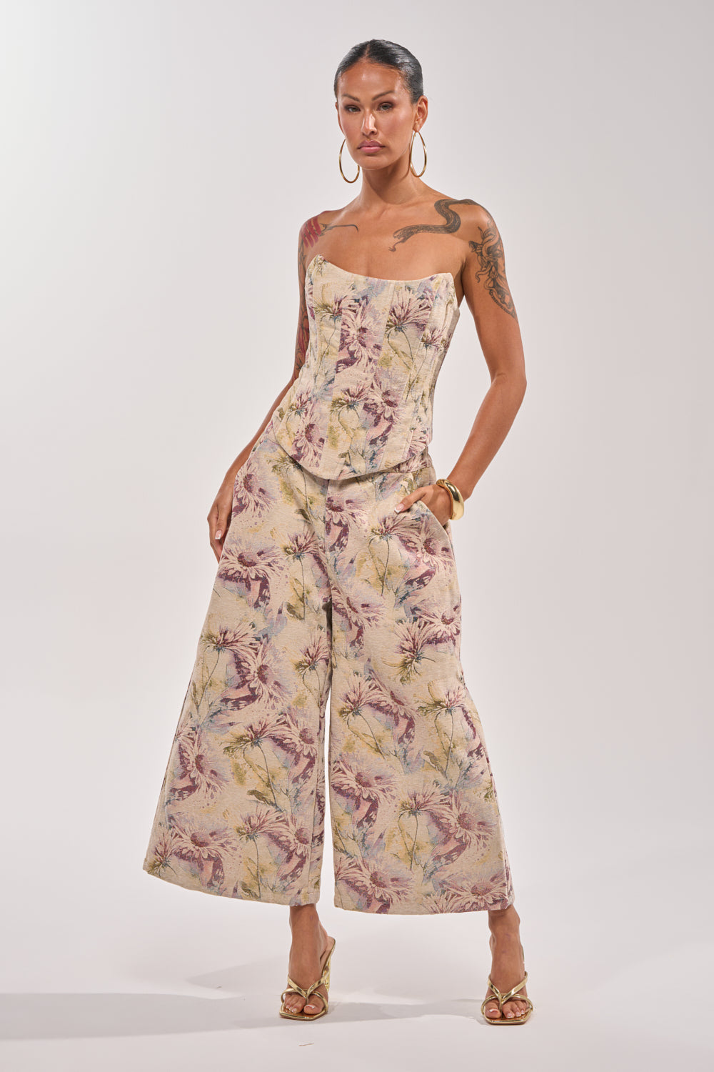A woman stands confidently against a plain background, wearing the SWEETIE PIE BROCADE PANT—a wide-leg, cropped style with a pastel floral and bird print—paired with a strapless fitted top, gold hoop earrings, and heeled sandals. 5