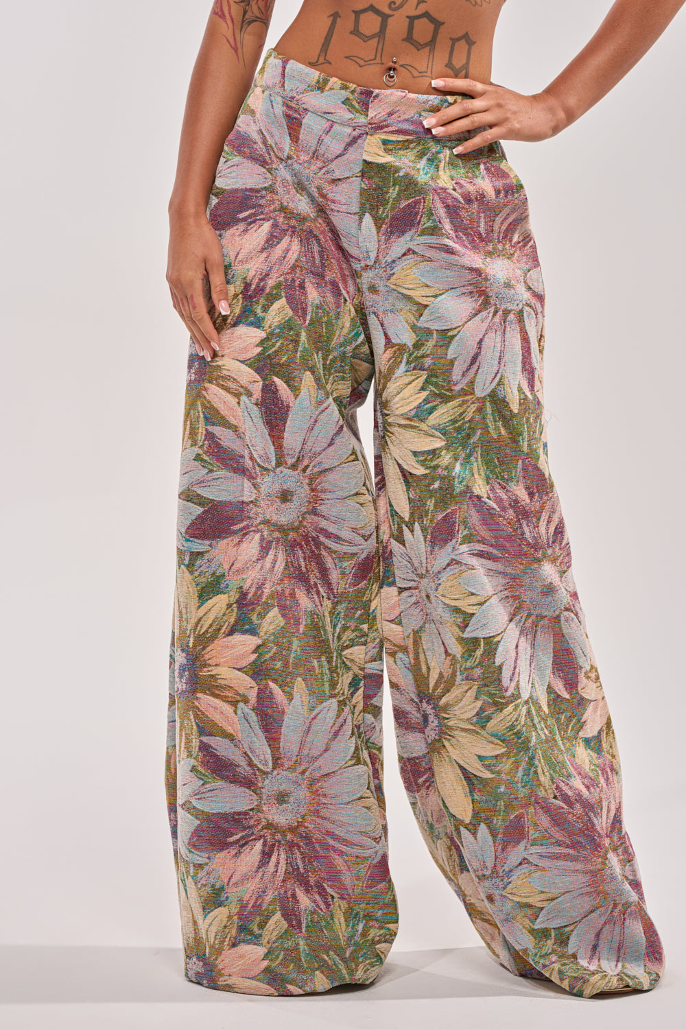 A person wearing the SUNFLOWER FIELDS WILD LEG PANT, featuring high-rise floral trousers with a bold, oversized print in purple, green, yellow, and blue. Their midriff is exposed, revealing a "1999" tattoo on their stomach. 1