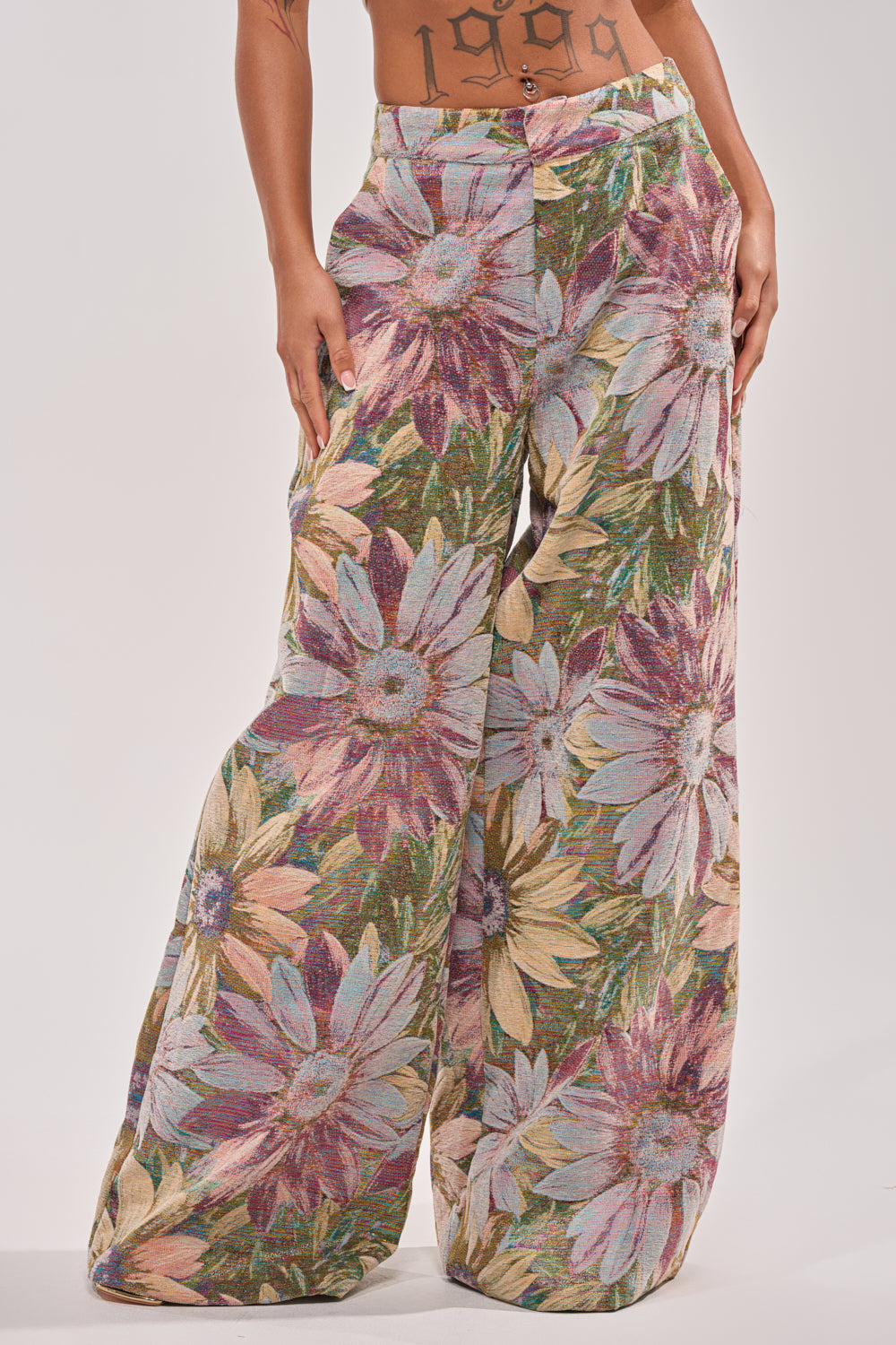 Wearing the SUNFLOWER FIELDS WILD LEG PANT, featuring a bold sunflower print in purple, yellow, green, and blue on textured fabric; midriff tattoos peek out above these high-rise statement trousers. 2