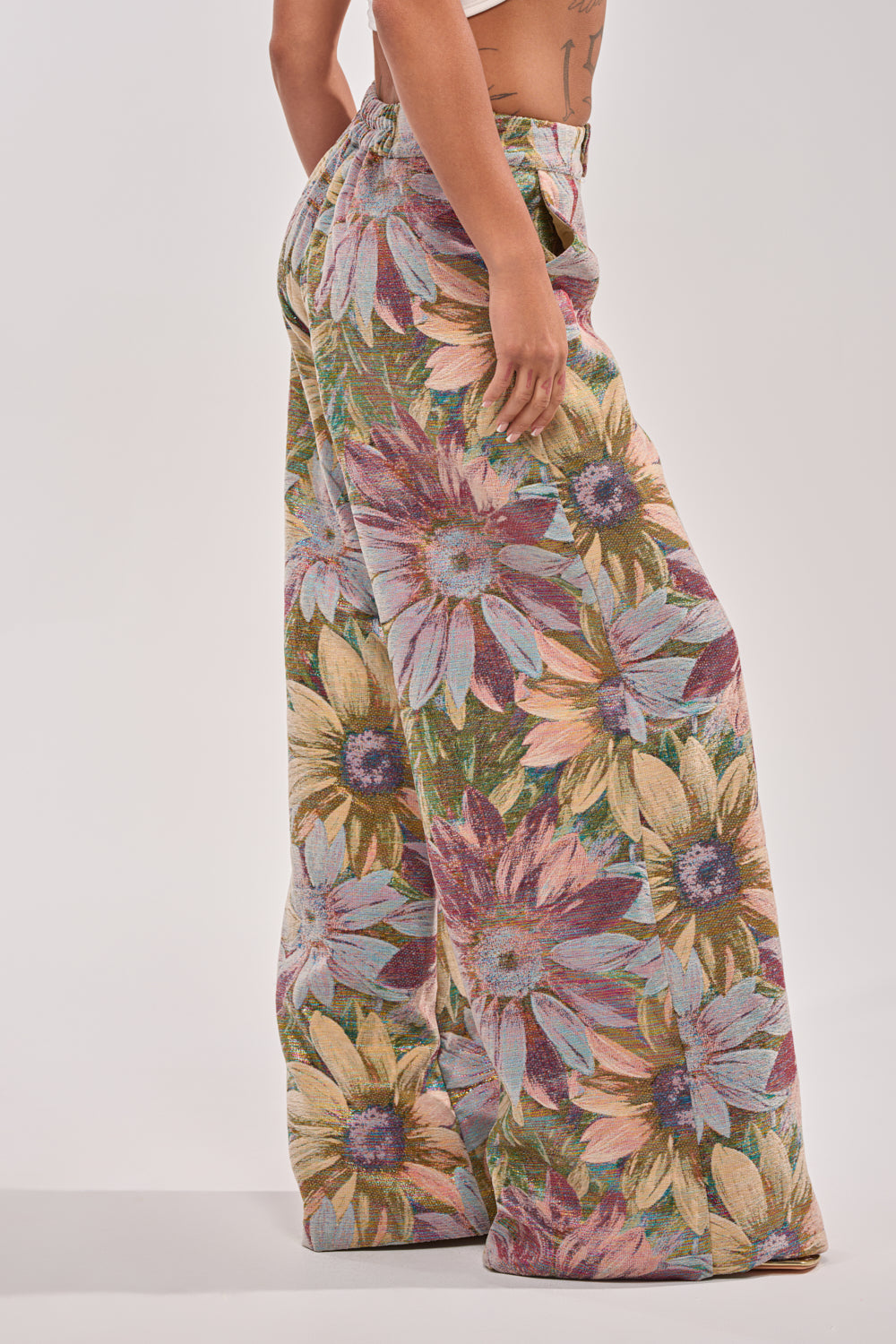 A model showcases the SUNFLOWER FIELDS WILD LEG PANT, featuring a high-rise waist and bold sunflower print in yellow, purple, and green, set against a plain light background. 3