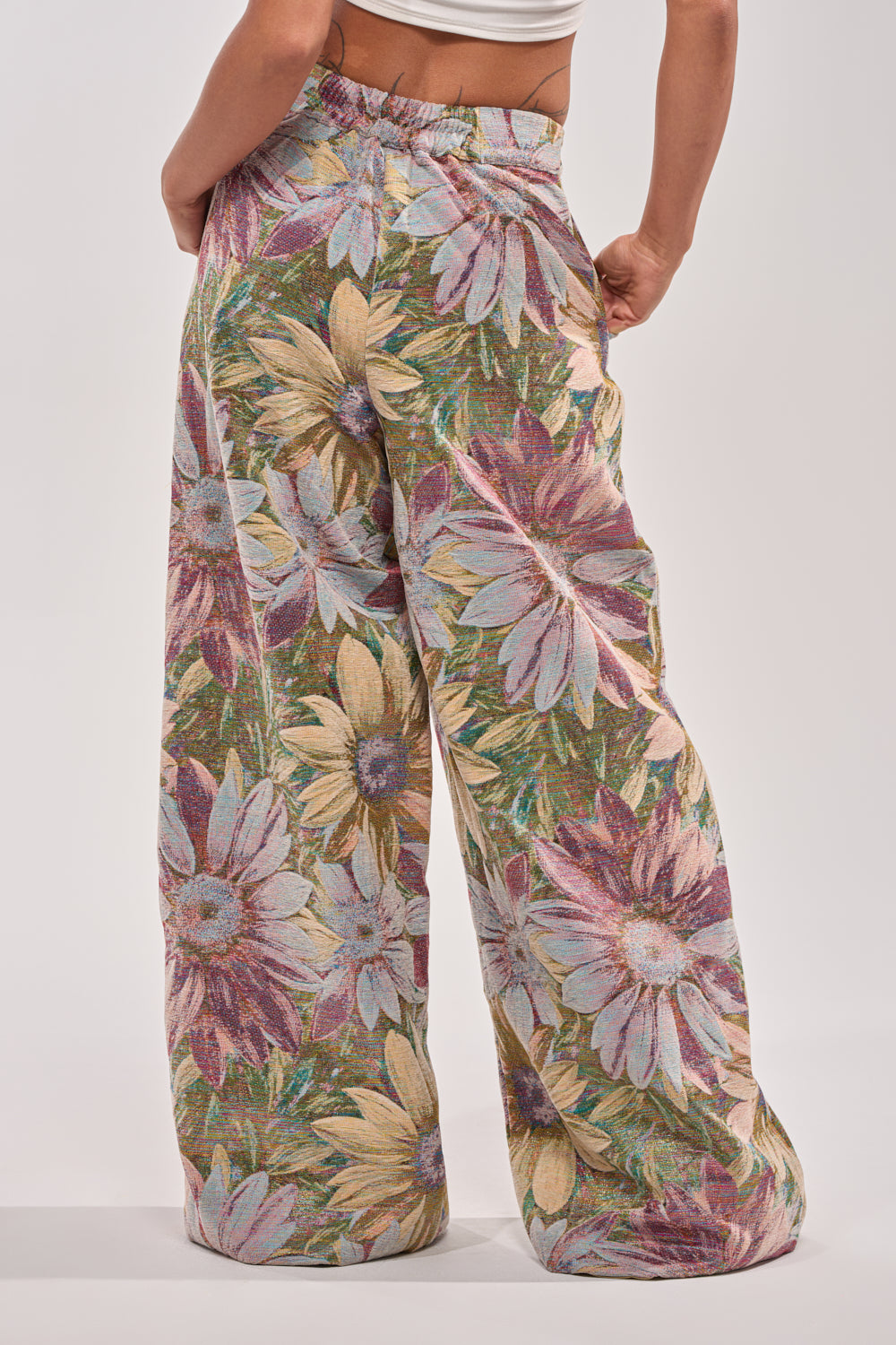 Someone stands with their back to the camera against a plain background, wearing SUNFLOWER FIELDS WILD LEG PANT featuring a vibrant sunflower print and high-rise fit. 4