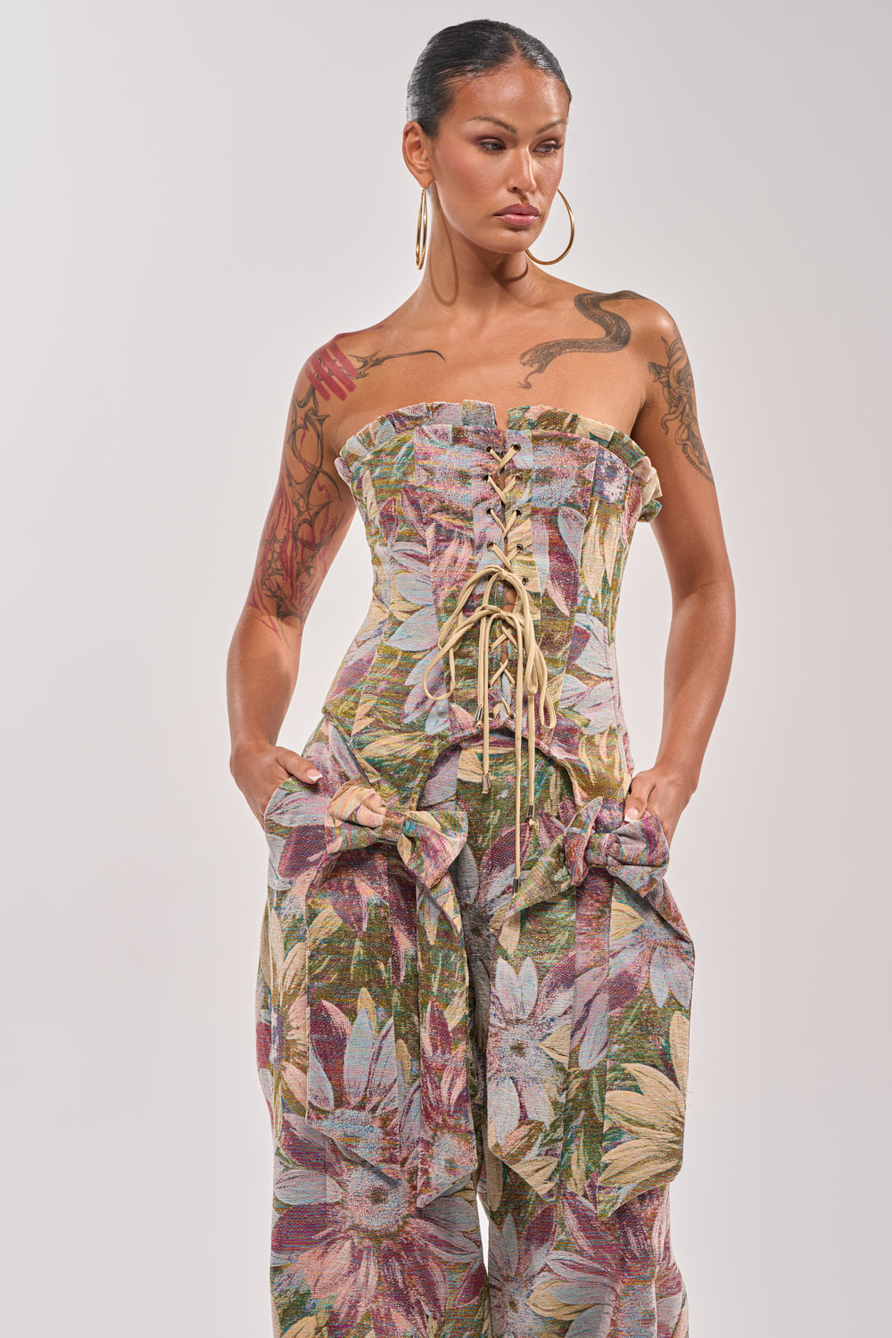 A woman with large hoop earrings and tattoos models the strapless, lace-up SUNFLOWER FIELDS CORSET velvet jumpsuit with her hands in the pockets, standing against a plain background. 0