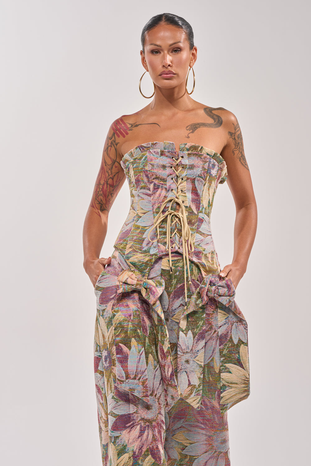 A woman with tattoos and hoop earrings confidently poses against a plain background, hands in the pockets of the strapless SUNFLOWER FIELDS CORSET dress, which features a metallic floral print in muted tones and elegant lace-up detailing. 1