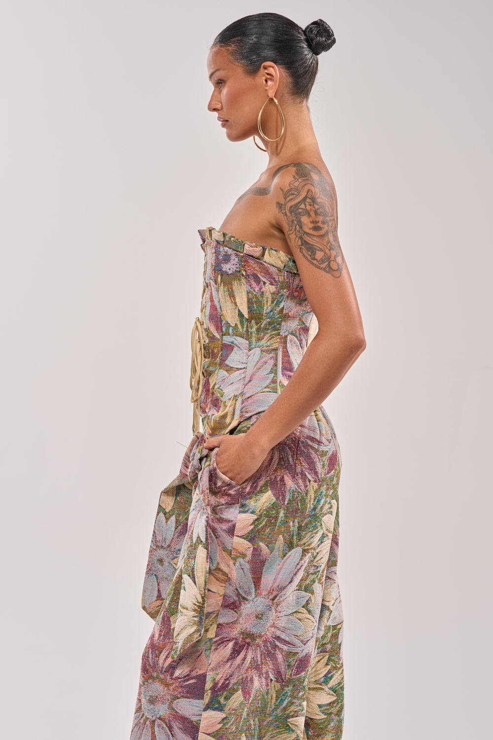 A woman in profile stands against a plain background, wearing the SUNFLOWER FIELDS CORSET—a strapless metallic floral print dress—with her hair in a bun, large hoop earrings, and a tattoo on her upper arm for party-ready style. 2