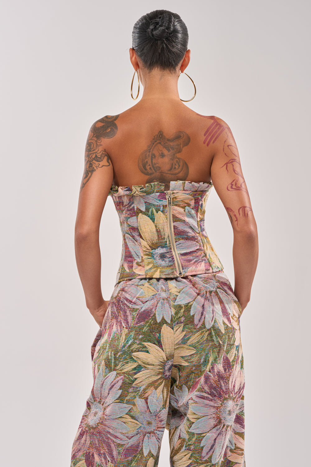 A person with large hoop earrings and visible tattoos wears the SUNFLOWER FIELDS CORSET, a strapless ruffle top paired with matching metallic floral wide-leg pants. They're shown from behind, hair styled in a neat bun. 3