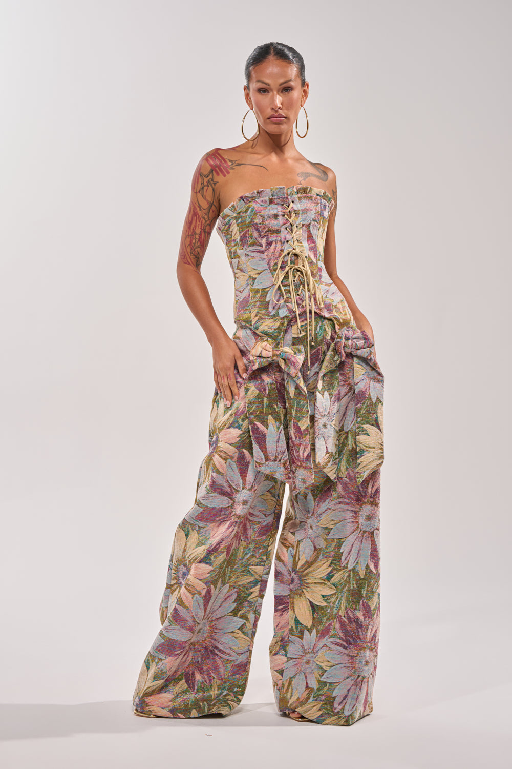 A person stands confidently against a plain background, wearing the SUNFLOWER FIELDS CORSET—a strapless, floral velvet top—paired with wide-leg metallic floral print pants, hoop earrings, and showing arm tattoos. 4