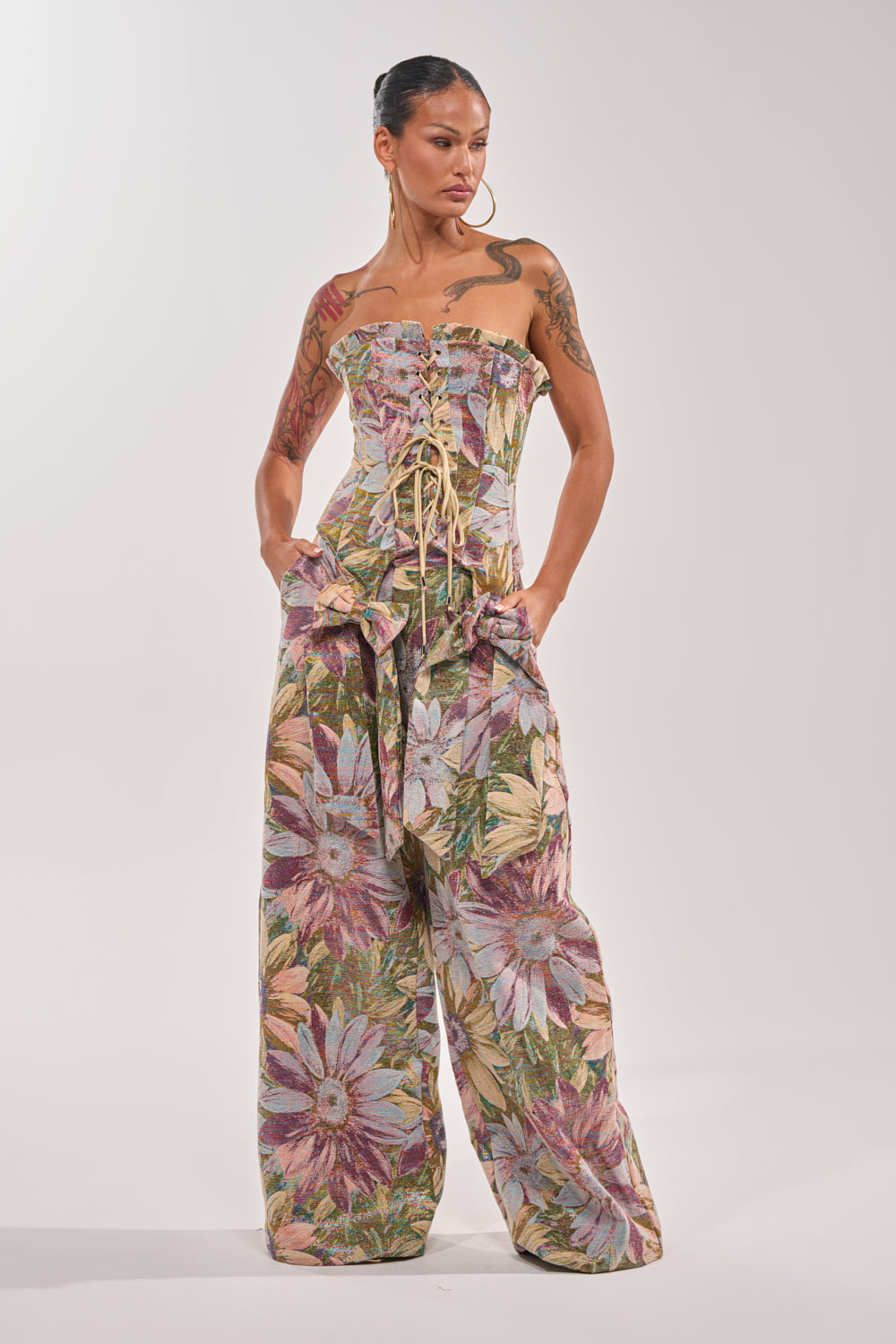 A woman with visible arm tattoos and her hair pulled back stands confidently wearing the SUNFLOWER FIELDS CORSET, featuring a metallic floral print, strapless design, lace-up front, and wide-leg jumpsuit styling. 5