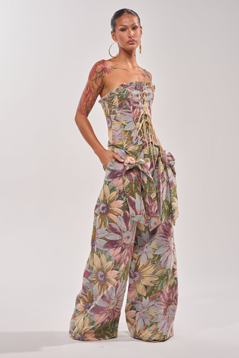 A woman wears the SUNFLOWER FIELDS CORSET, a strapless, lace-up jumpsuit with wide legs and a metallic floral print. Hoop earrings, visible tattoos, and hands in pockets complete her party-ready look against a plain background. 6