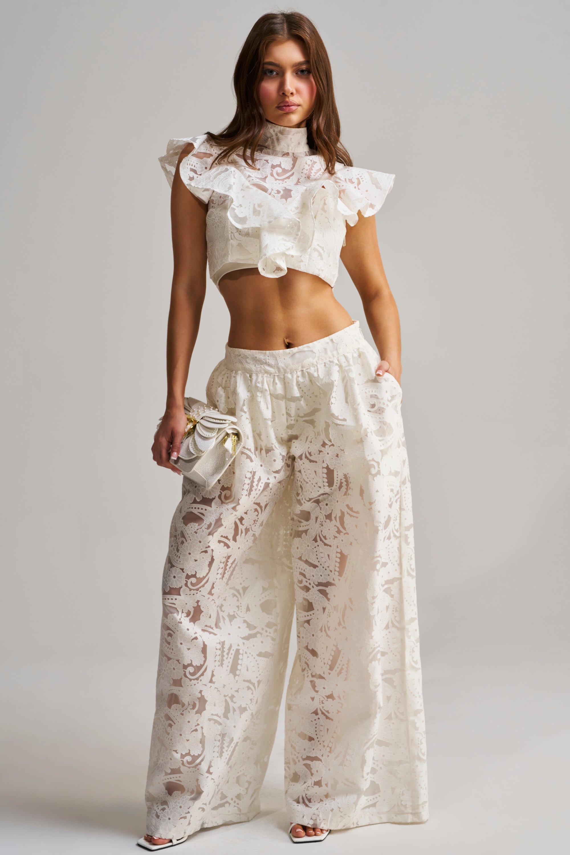 A woman poses in ivory floral lace EMMA EMBROIDERED BALLOON PANT and a ruffled crop top, holding a small purse. She completes her spring look with white heels, set against a plain light grey background.