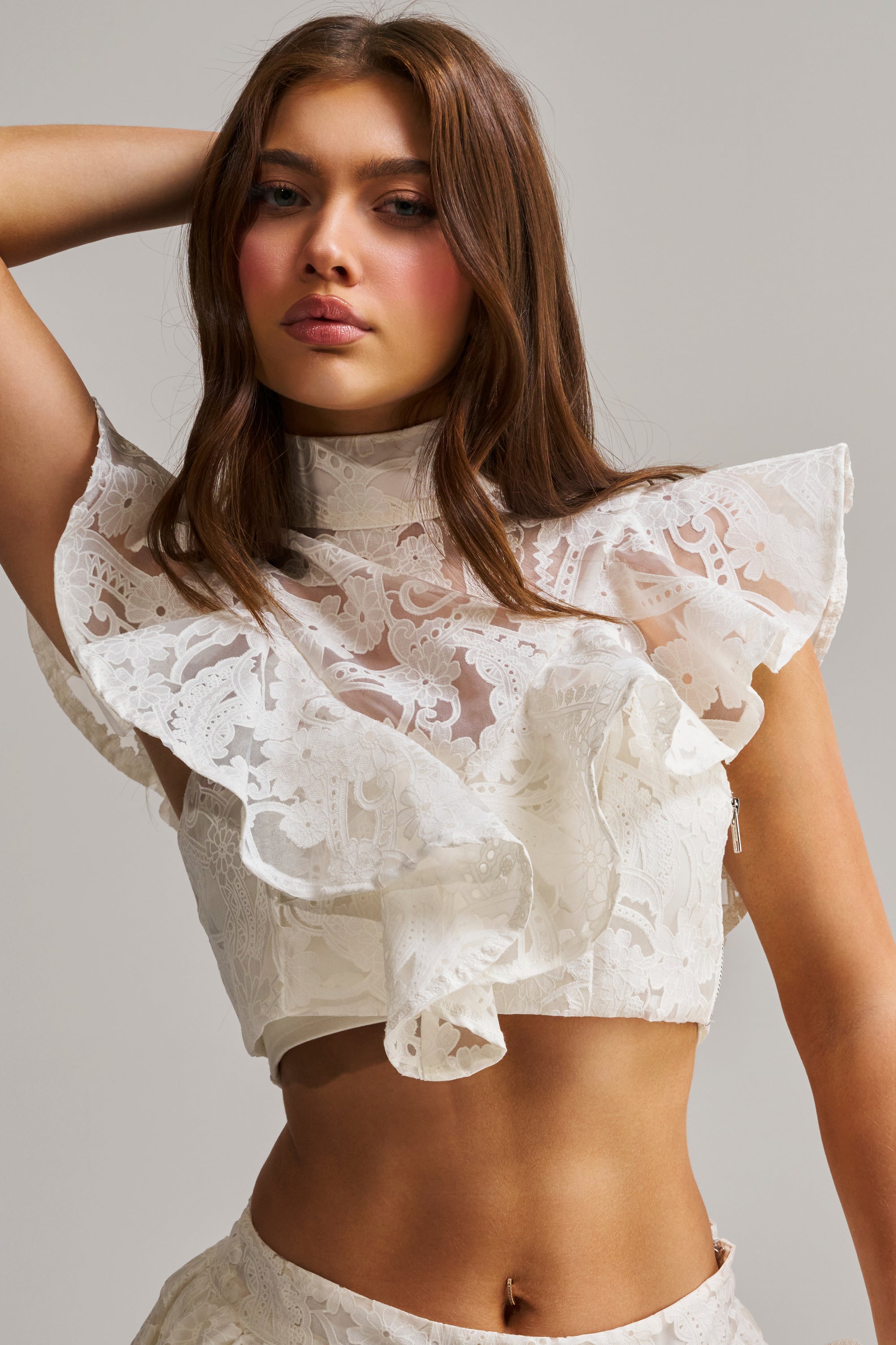 A woman with long brown hair poses confidently in the EMMA EMBROIDERED CROPPED BLOUSE, a white mockneck top with ruffle and lacy sleeveless details, standing against a plain light gray background.