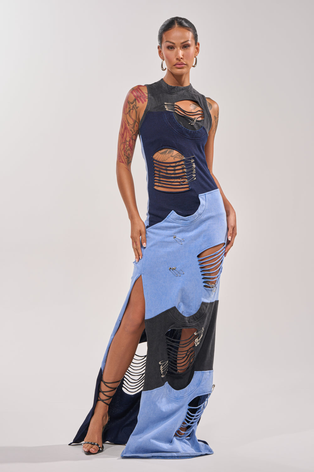 A woman exudes confidence in the LAST MINUTE MASTERPIECE DISTRESSED MAXI DRESS, featuring bold cutouts, safety pins, and shredded details. She pairs it with strappy heels and shows off her tattoos against a plain light background. 0