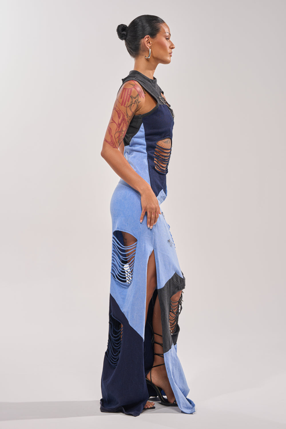 A woman with her hair in a bun stands in profile, wearing the LAST MINUTE MASTERPIECE DISTRESSED MAXI DRESS—sleeveless with slits and distressed panels—paired with large hoop earrings, visible arm tattoos, and black heels. 3