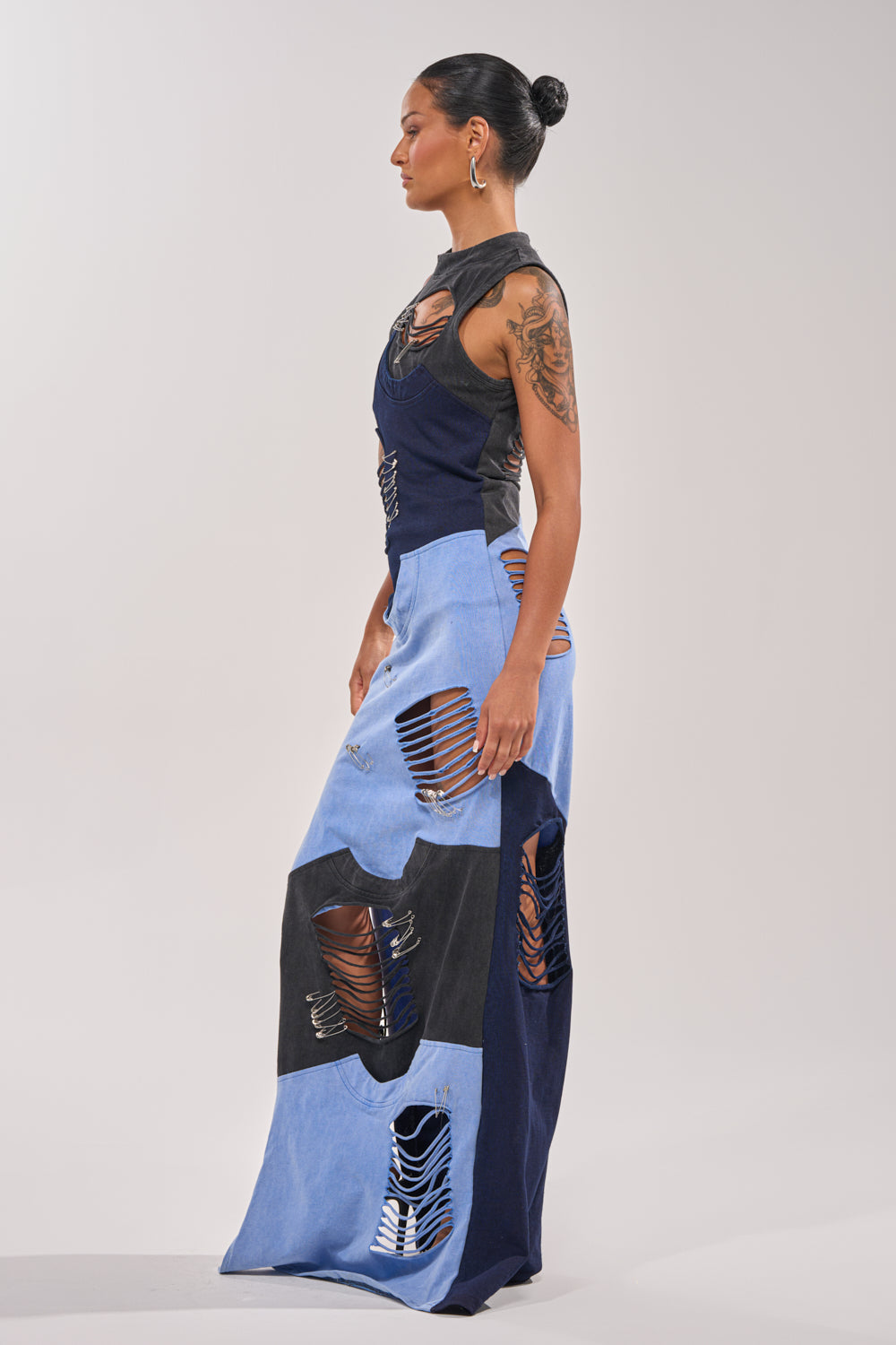 A woman with a tattooed arm and hair in a bun models the LAST MINUTE MASTERPIECE DISTRESSED MAXI DRESS, a form-fitting color block maxi with patchwork denim panels, distressed details, and bold horizontal cut-outs, standing in profile against a plain background. 4