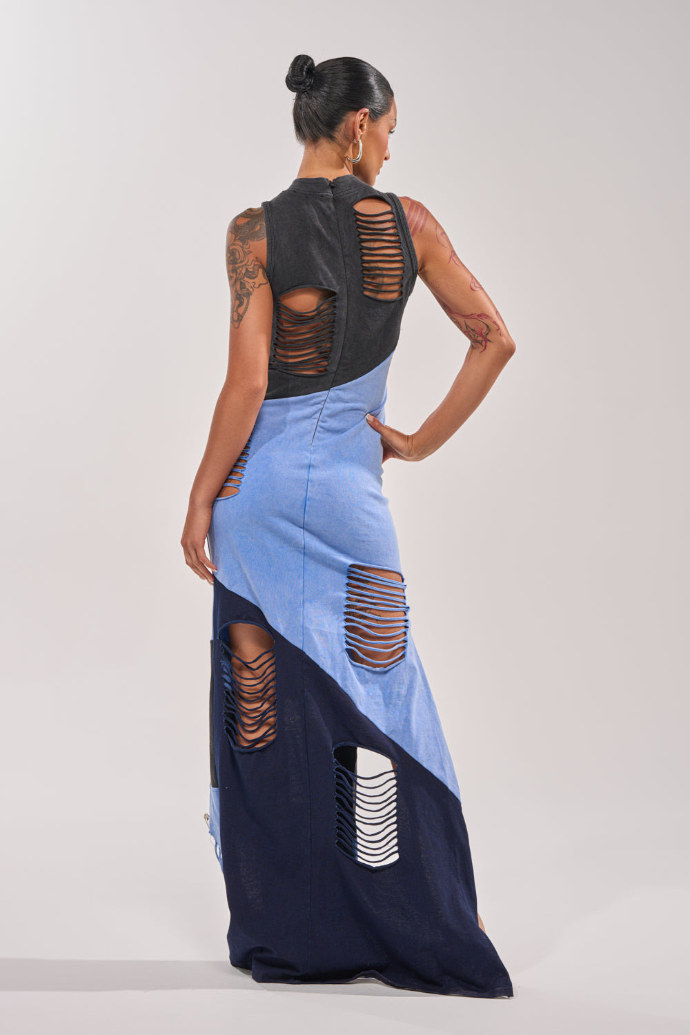 A woman faces away, wearing the LAST MINUTE MASTERPIECE DISTRESSED MAXI DRESS in black and blue color block with multiple horizontal cutouts. Her hair is in a bun and tattoos are visible on her arms. 5