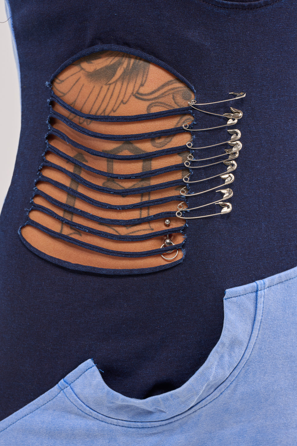 Close-up of the LAST MINUTE MASTERPIECE DISTRESSED MAXI DRESS in navy with distressed paneling, safety pin details, and horizontal cutouts revealing tattooed skin. A light blue shirt overlaps at the bottom with a semi-circular cutout edge. 6