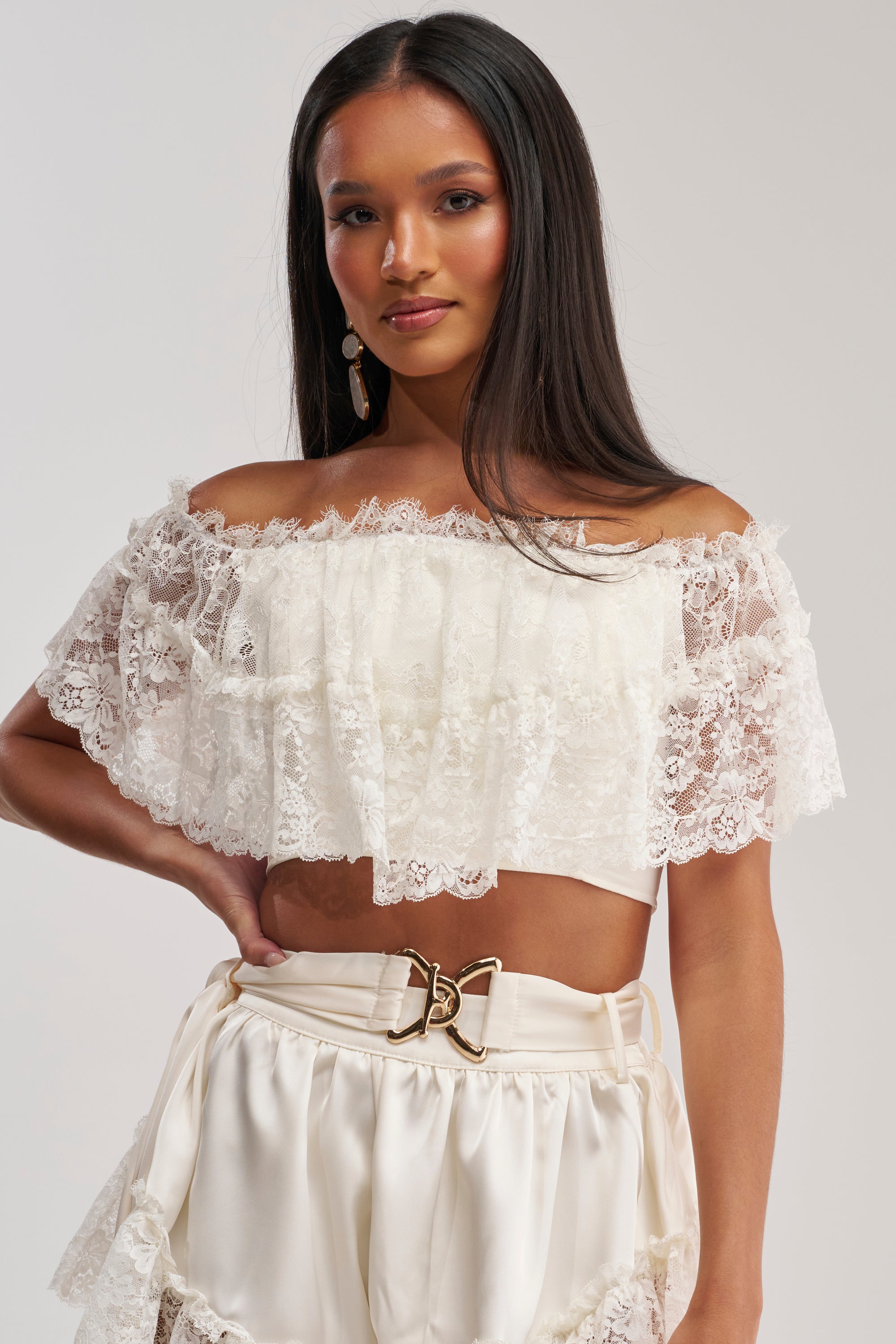 Channeling boho-chic style, a woman with long dark hair wears the PRIMROSE LACE BLOUSE and high-waisted cream shorts with a gold belt, standing against a plain background.