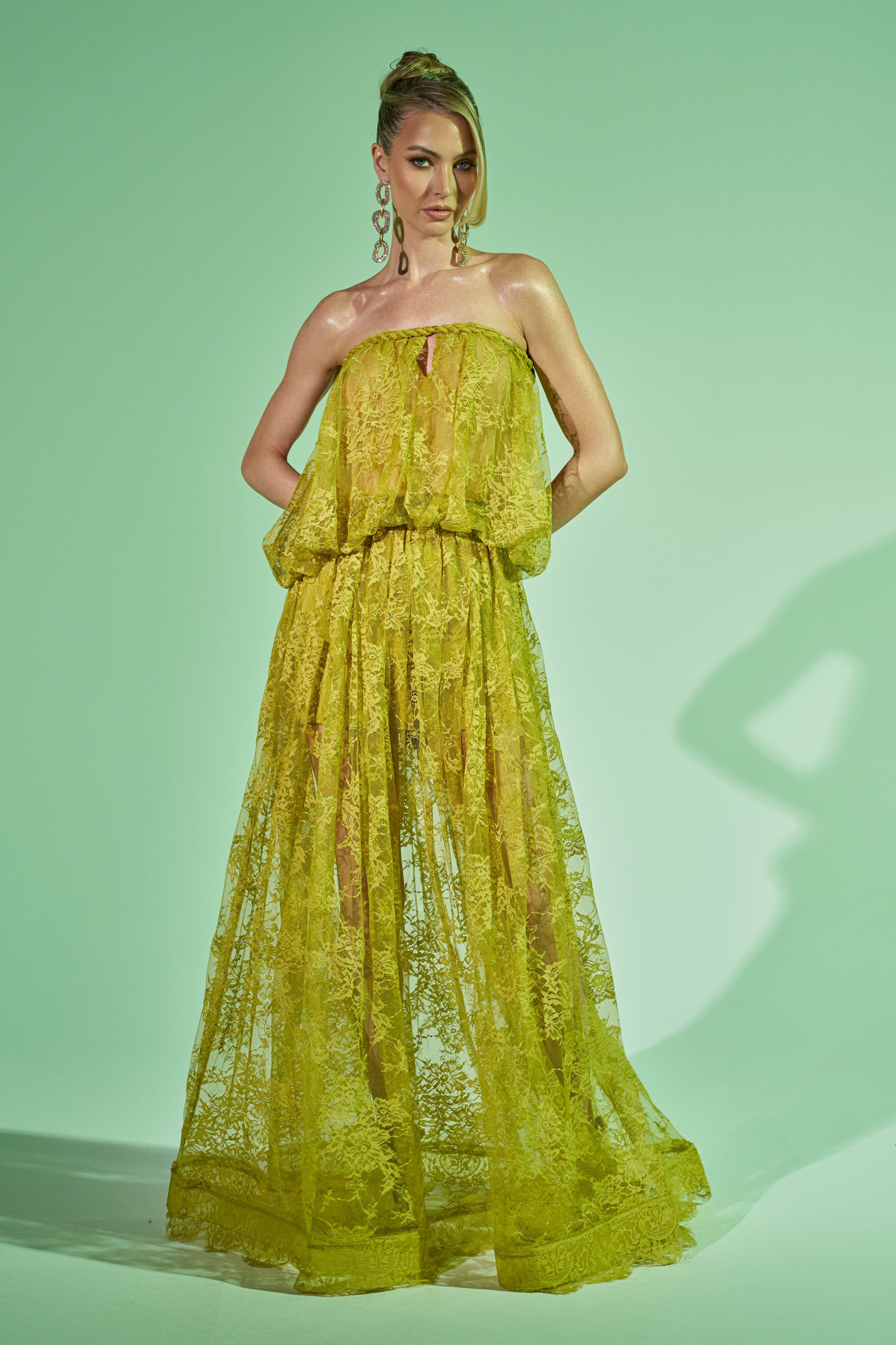 A woman stands against a light green backdrop wearing the HANNAH LACE MAXI SKIRT, a strapless yellow lace dress with a flowing sheer maxi skirt. She has her hands behind her back, wears large earrings, and her hair is styled up. 0