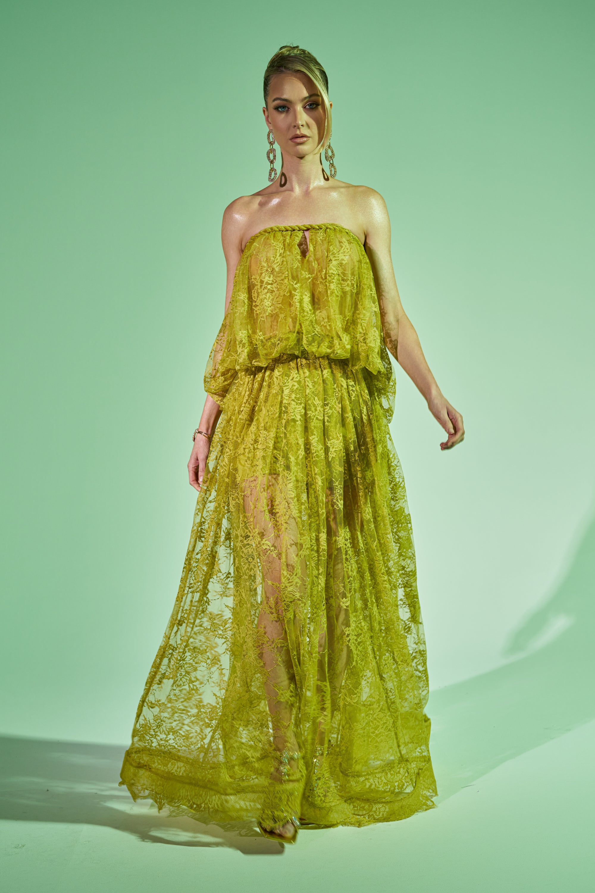 A woman poses against a green backdrop wearing the HANNAH LACE MAXI SKIRT, a strapless, sheer yellow lace maxi dress with a flowing skirt. She accessorizes with large dangling earrings and has her hair styled up. 1