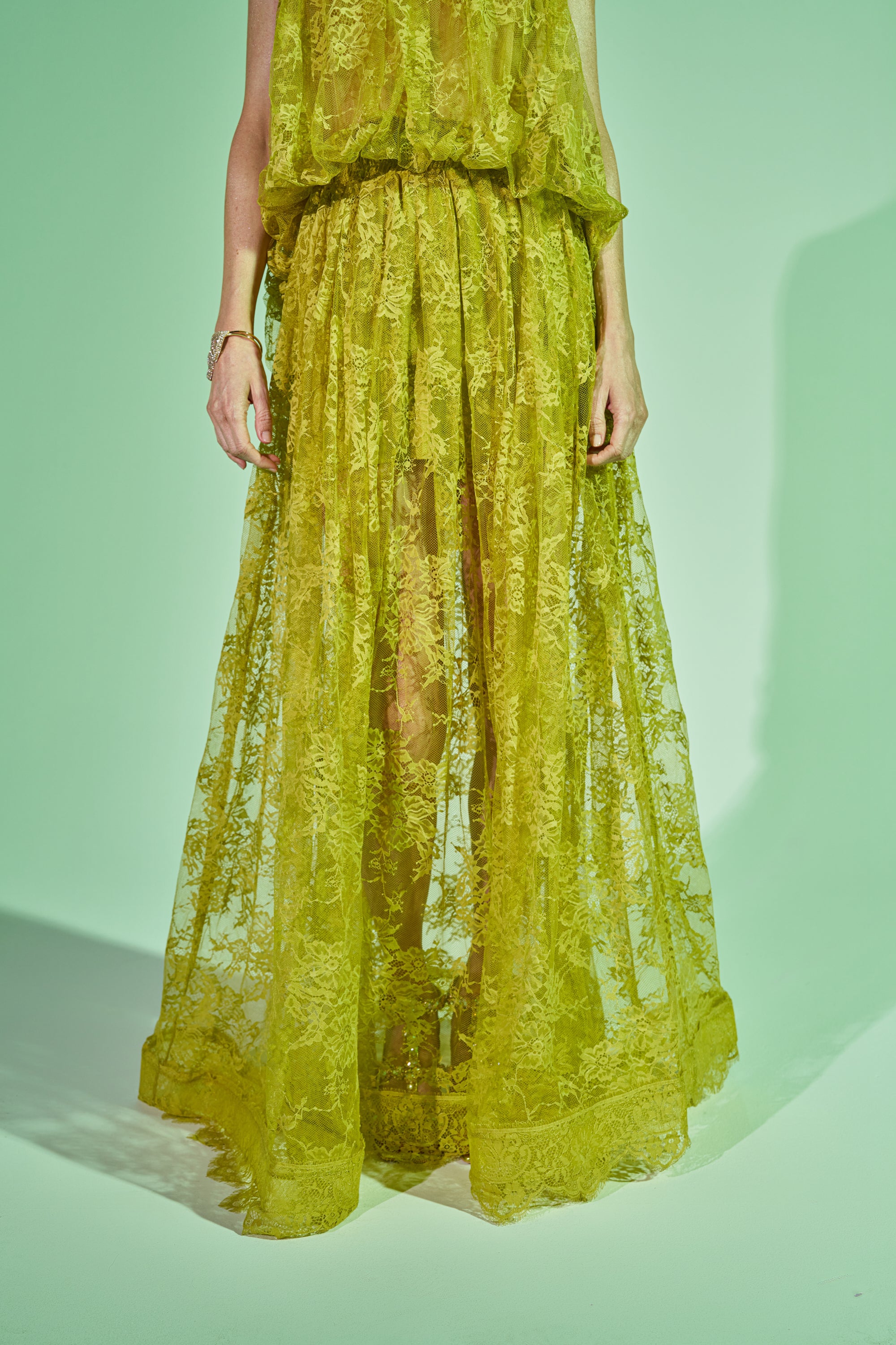 Wearing the HANNAH LACE MAXI SKIRT in sheer, bright yellow chartreuse, a person stands against a light green background. The floor-length lace skirt is sleeveless and reveals the outline of their legs underneath. 2