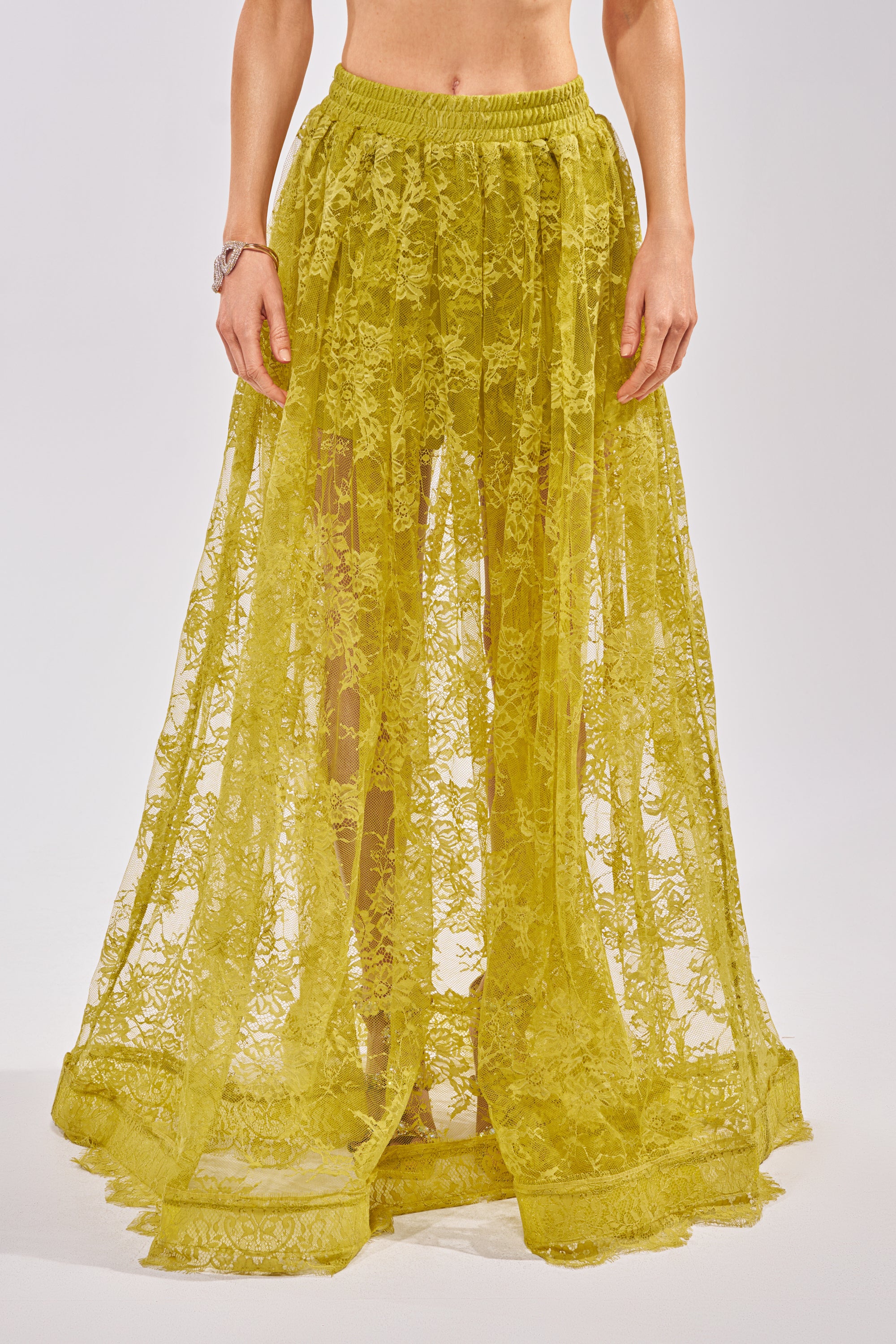A person wears the HANNAH LACE MAXI SKIRT—a yellow, sheer lace maxi skirt with a gathered waistband. The see-through design reveals their legs as they stand against a plain light background. 3