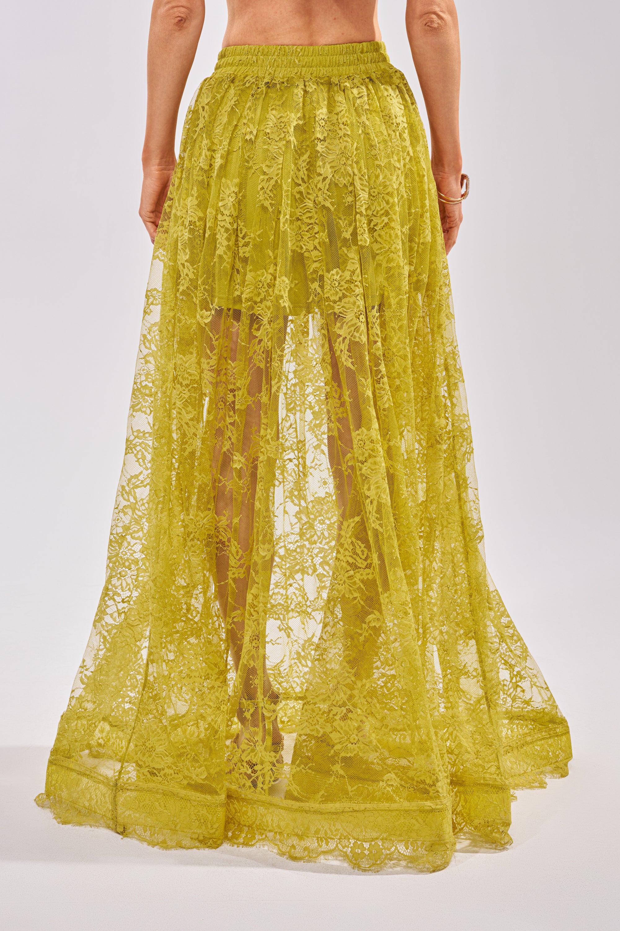 A person models the HANNAH LACE MAXI SKIRT—a long, sheer chartreuse lace design with a visible waistband—against a plain light background. The transparent, flowy skirt elegantly reveals the legs underneath. 5