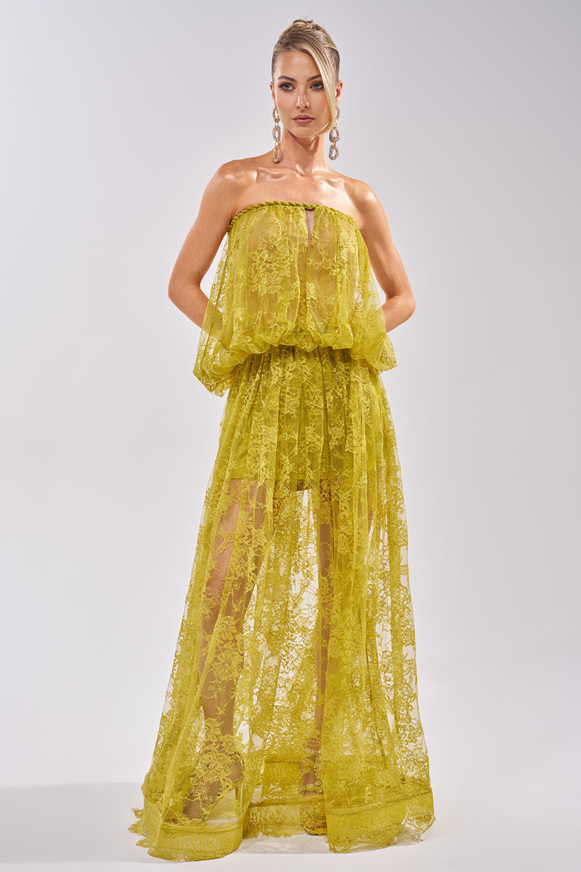 A woman exudes confidence in the HANNAH LACE MAXI SKIRT, featuring bright yellow lace with a sheer maxi silhouette and layered bodice. She pairs it with gold earrings and an updo, standing against a light background for a fresh spring look. 7