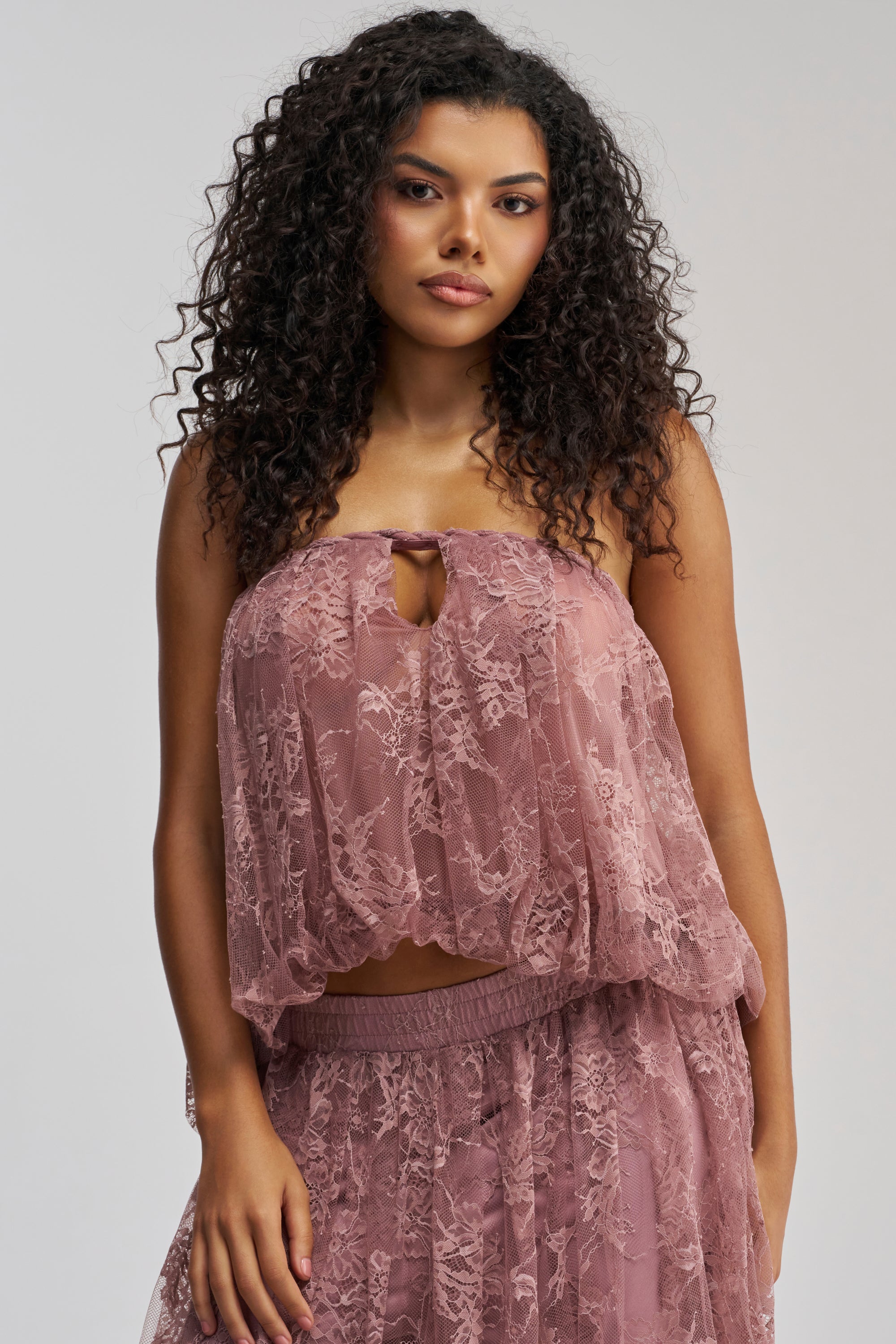 A woman with curly hair stands confidently against a neutral background, wearing the HANNAH LACE BLOUSE IN MAUVE—an elegant strapless top that’s perfect for making a statement on date night.