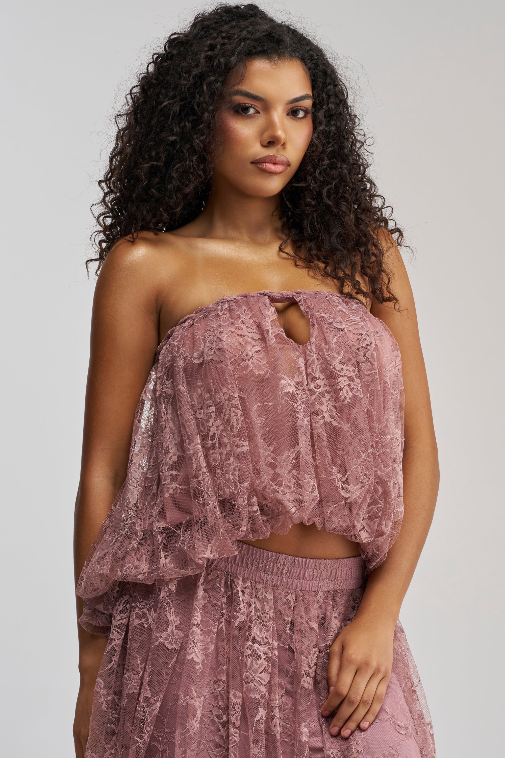 A woman with curly hair wears the HANNAH LACE BLOUSE IN MAUVE, styled strapless with a matching skirt. She stands against a plain background, looking confidently at the camera—perfectly dressed for a chic date night look.