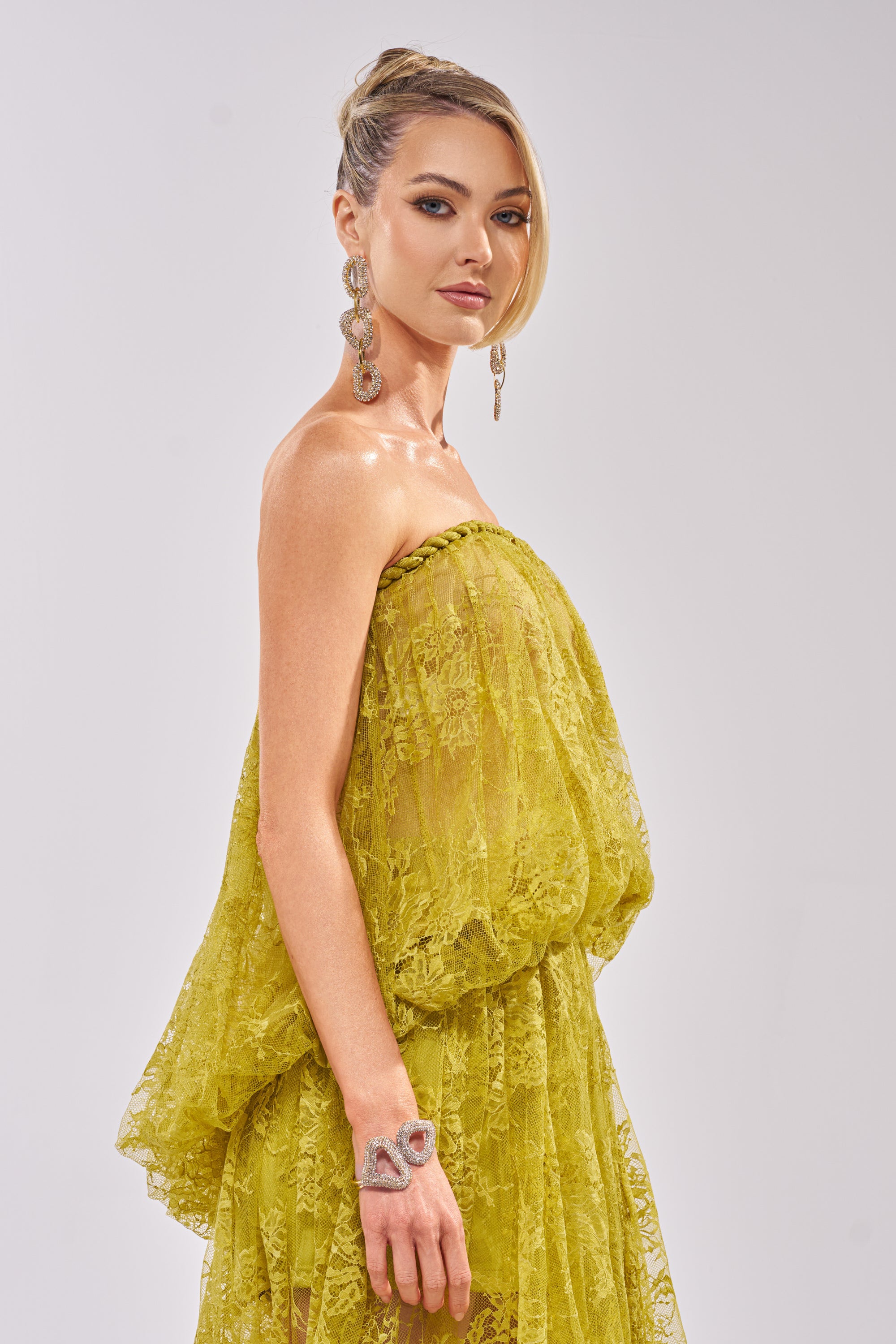 A woman poses sideways in the HANNAH LACE BLOUSE, a strapless sheer yellow lace top. She has blonde hair styled up, large dangling earrings, and a statement bracelet, standing against a plain light backdrop. 4